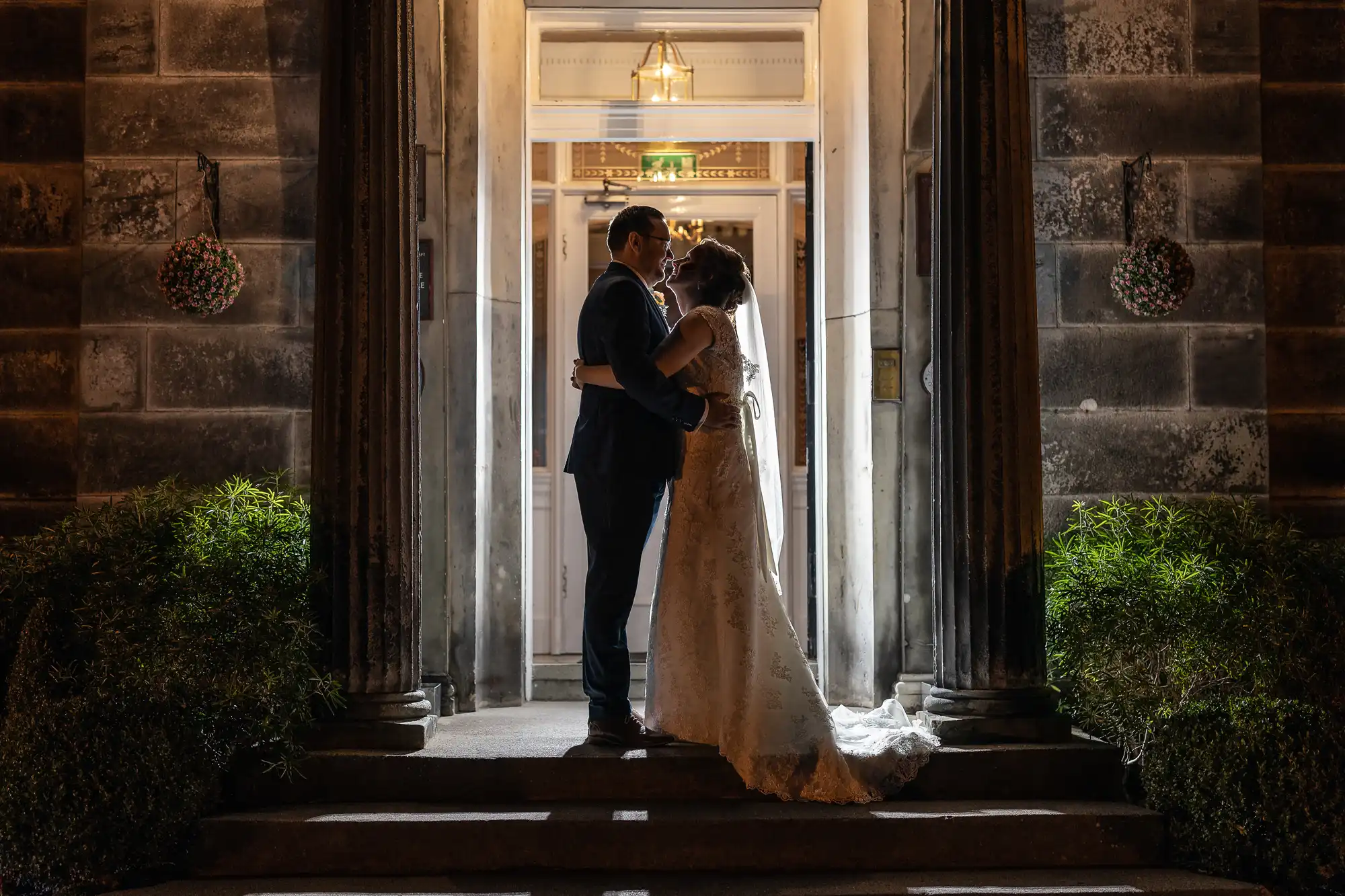 A bride and groom stand embracing and facing each other in the doorway of a grand building at night. They are silhouetted by warm indoor lights, surrounded by tall stone columns and plants on either side of the entrance.