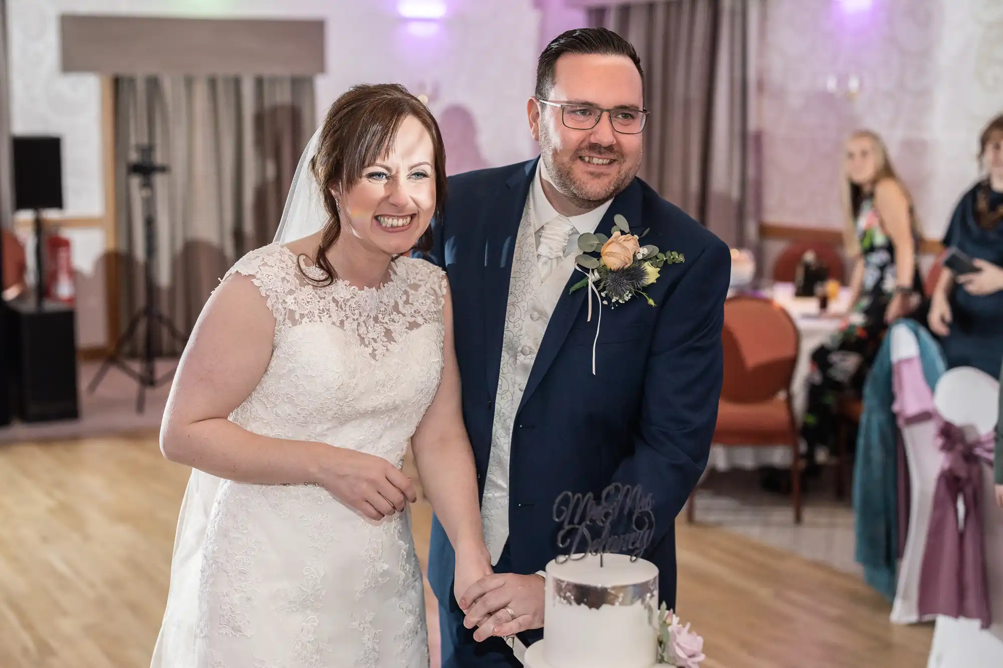 A smiling bride in a white lace dress and a groom in a navy suit with a buttonhole cut a white wedding cake together indoors, surrounded by decorated tables and guests in the background.