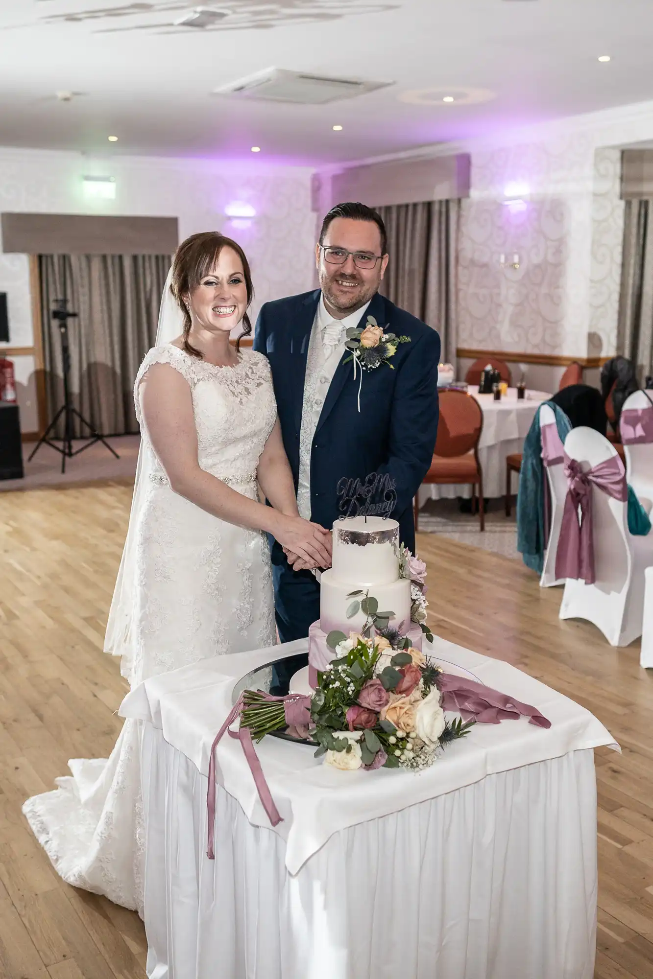 A bride in a white lace gown and a groom in a blue suit stand smiling, cutting a white wedding cake together. The cake sits on a white table adorned with a bouquet of flowers and pink ribbons in a decorated reception hall.