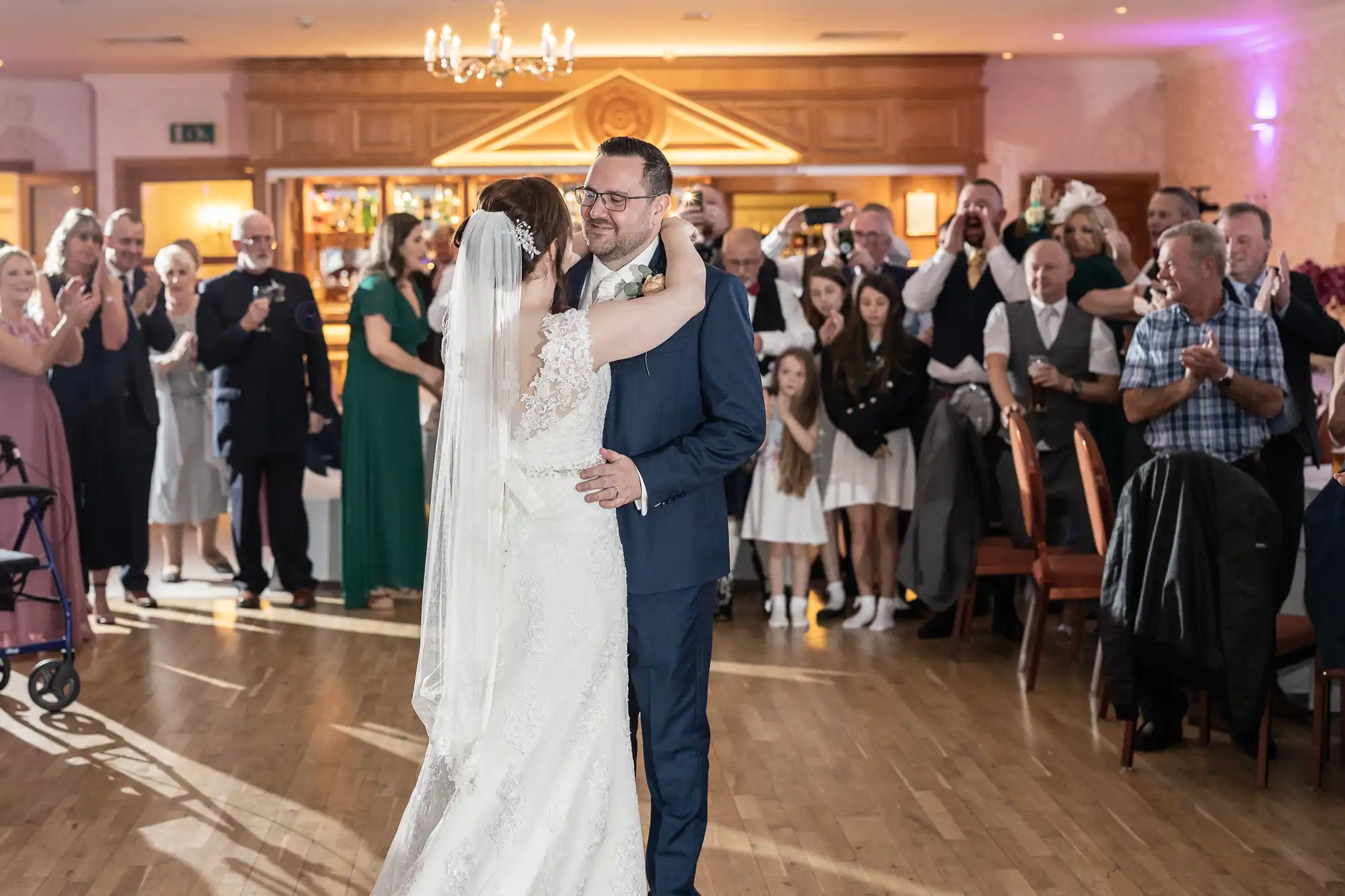 A bride and groom share their first dance in a brightly lit reception hall. The bride wears a white gown with lace sleeves and a veil; the groom wears a navy suit. Guests surround them, clapping and smiling in celebration. Warm lighting and wooden accents fill the room.