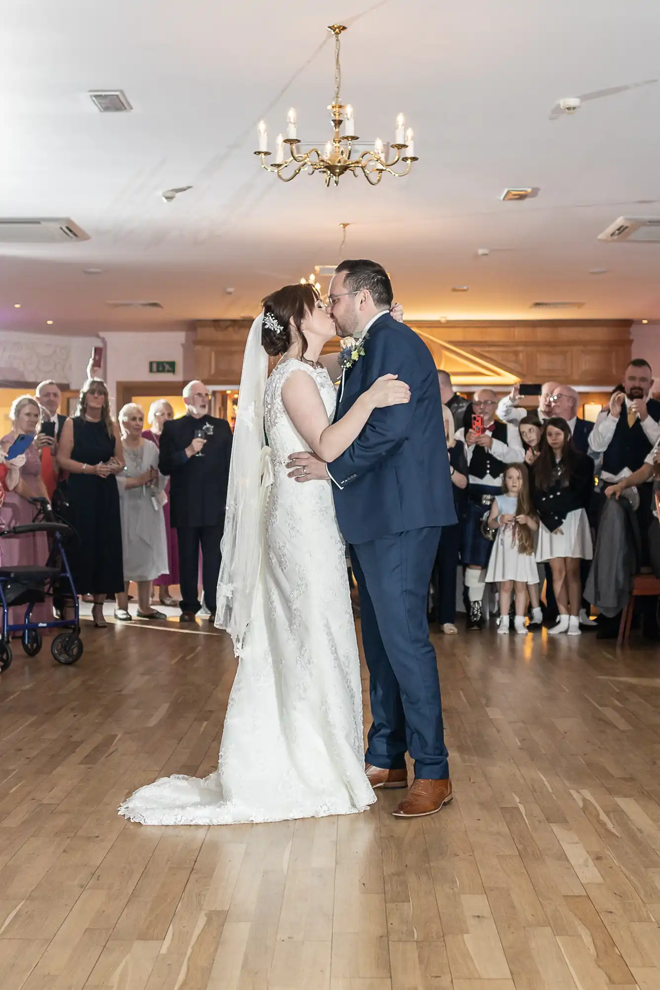 A bride in a white dress and veil and a groom in a blue suit share a kiss on a dance floor. Guests in formal attire stand around them, watching and taking photos. The room has wooden floors, a chandelier, and soft, warm lighting.