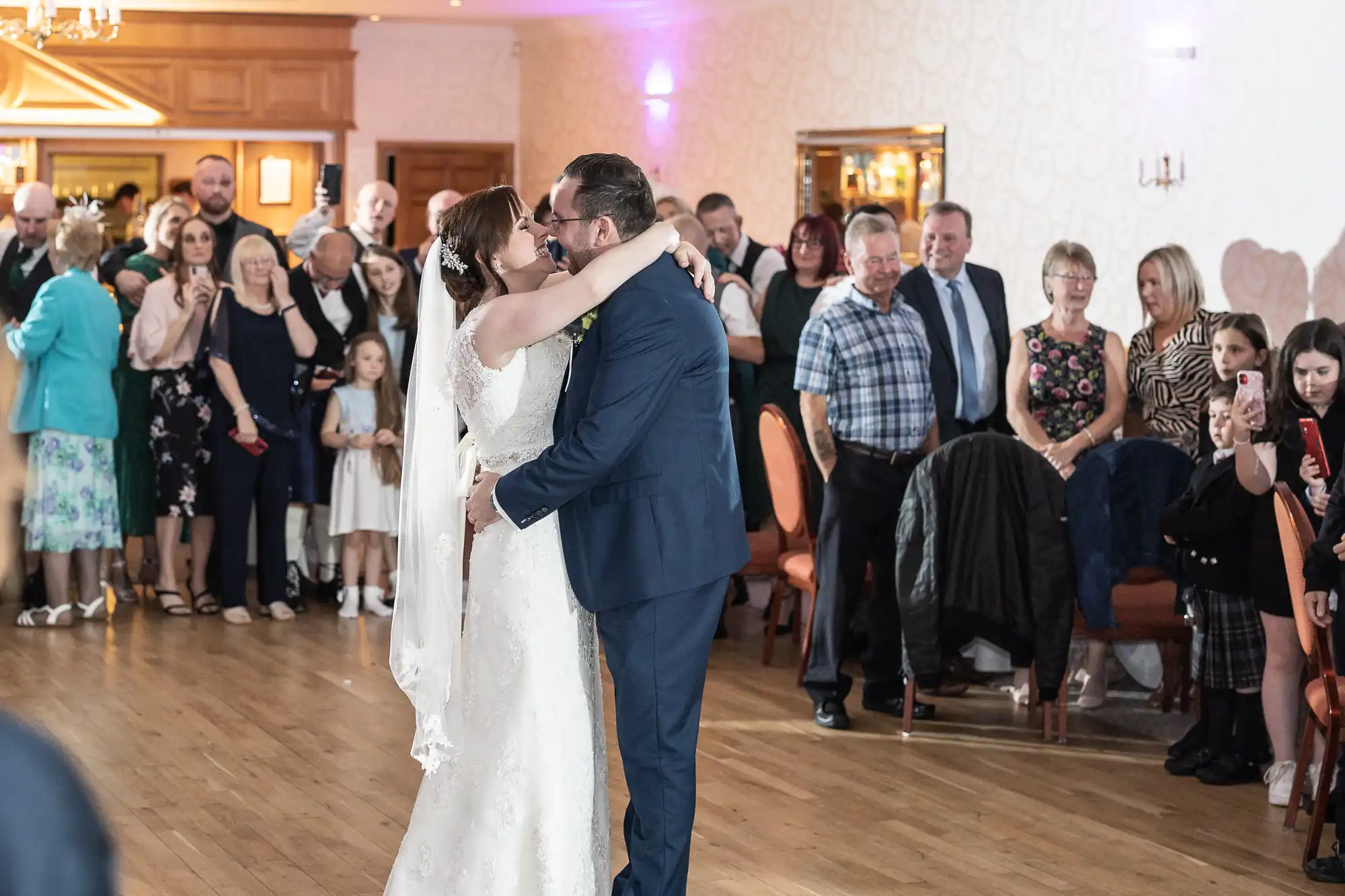 A bride and groom share a kiss on the dance floor during their wedding, surrounded by smiling guests. The bride wears a white dress and veil, and the groom wears a blue suit. Guests watch in the background, some seated, others standing, in a warmly lit venue.