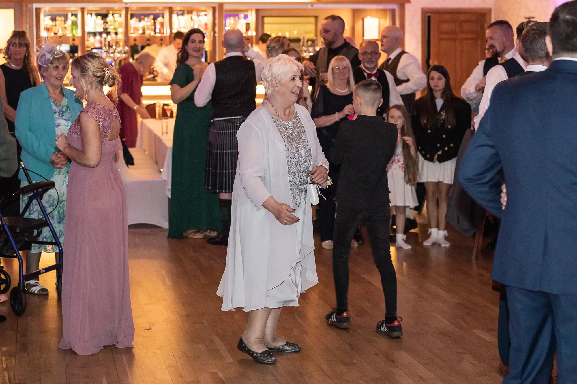 A group of people, young and old, gather in a warmly lit hall. A smiling elderly woman in a pale dress dances in the centre. Others stand, mingle, or watch, with a bar and rows of bottles visible in the background. The scene is lively and festive.