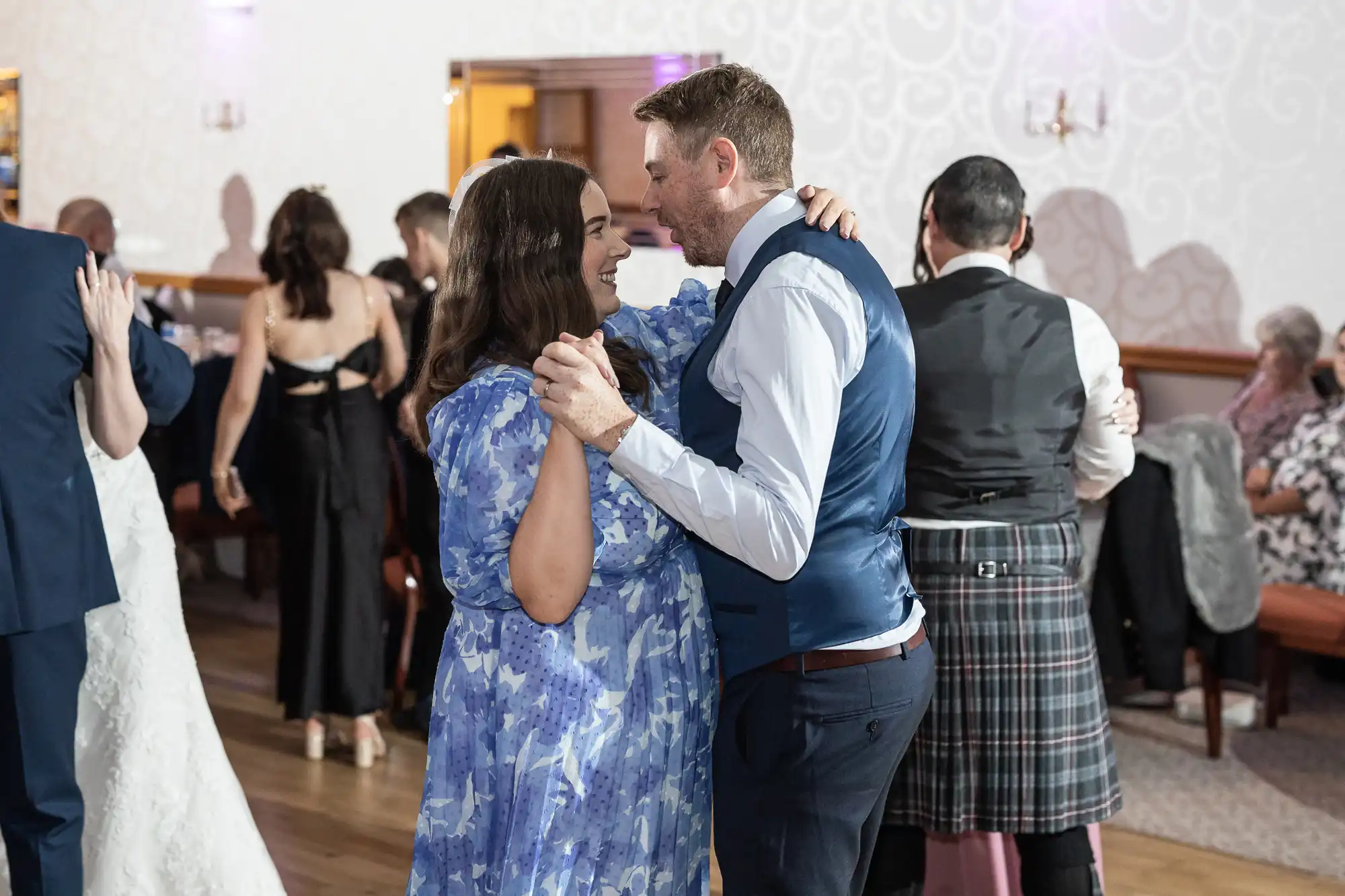 A man in a blue waistcoat and white shirt dances closely with a woman in a blue floral dress at a formal indoor event. Other couples dance in the background, including a man in a kilt. The room has white patterned walls and wooden floors.