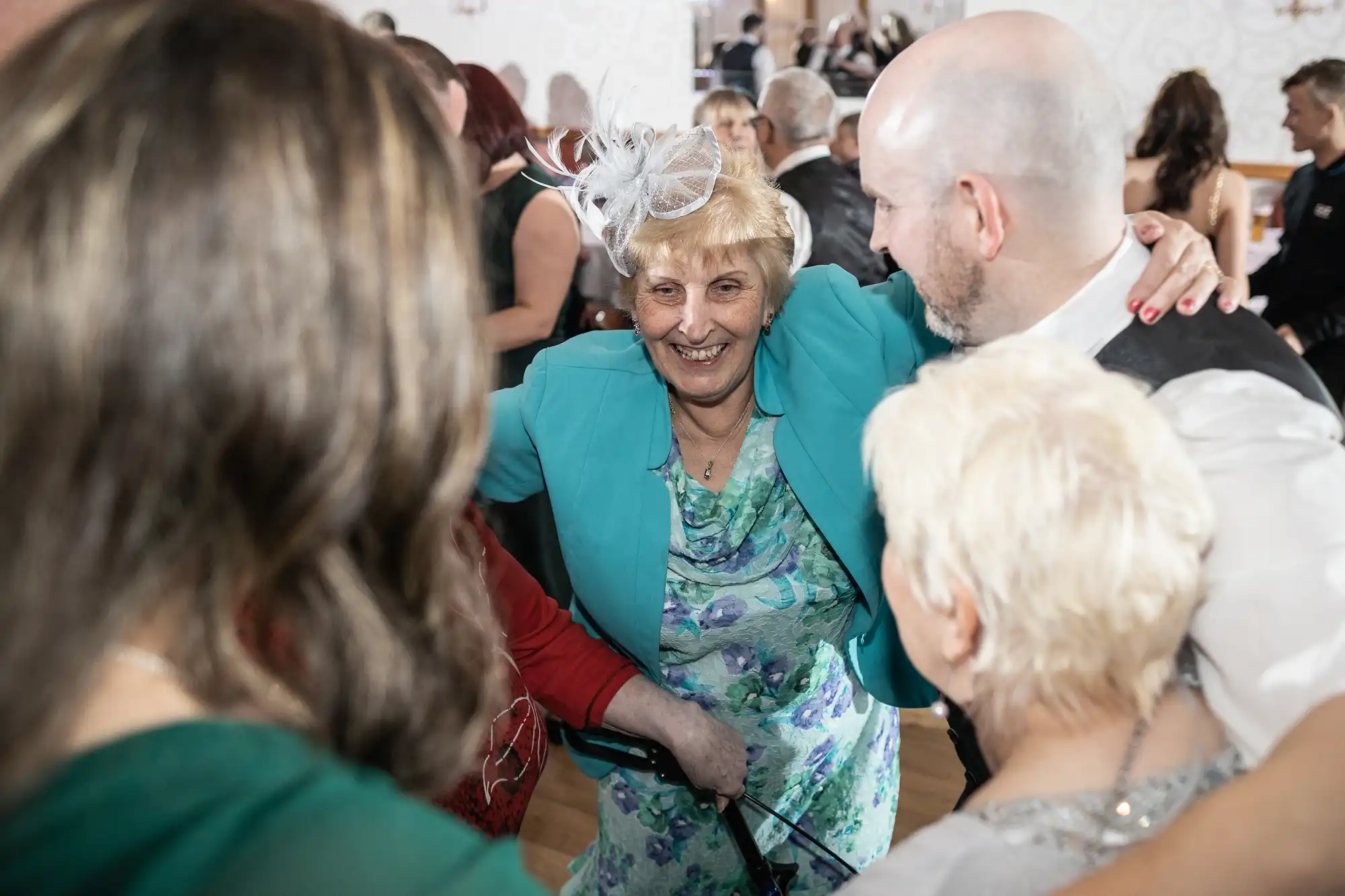 An elderly woman in a turquoise jacket and floral dress smiles warmly with arms around a group of people at a lively indoor event. The group, seen mostly from behind, gathers closely, suggesting celebration and joy. Other guests are visible in the blurred background.