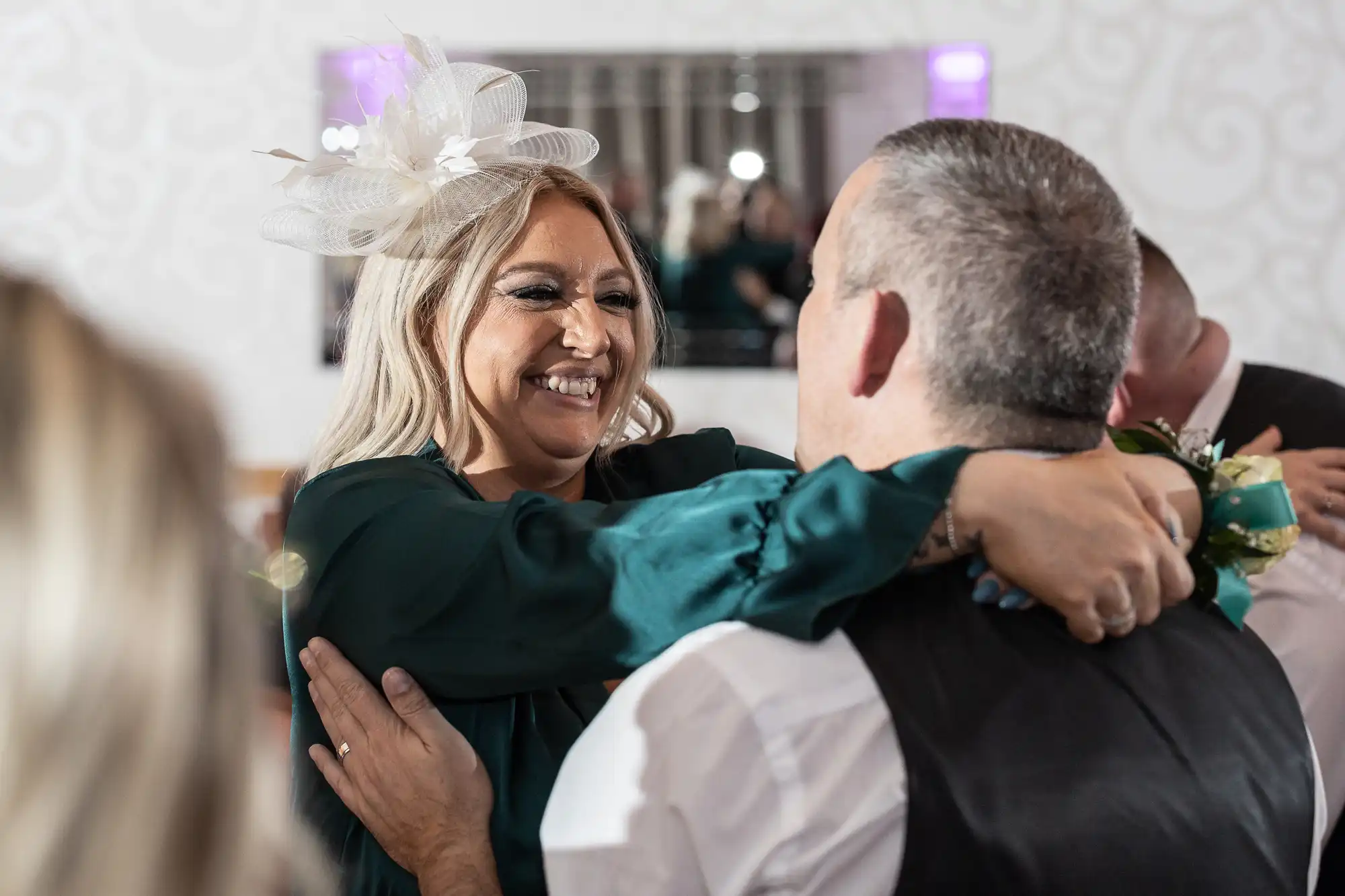 A smiling woman wearing a large white fascinator and green dress embraces a man in a white shirt and dark waistcoat. She looks happy, with her arms around his shoulders. People and a mirror appear in the softly lit, decorated background.