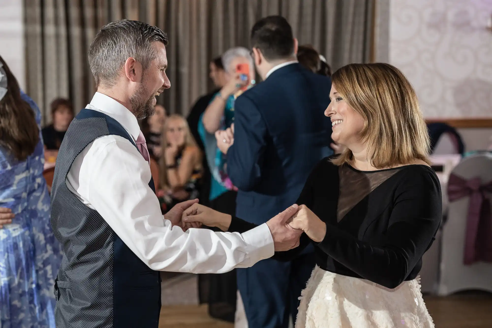 A smiling man in a waistcoat and tie holds hands and dances with a smiling woman in a black top and white skirt at an indoor event, with seated and standing guests in the background. The atmosphere appears joyful and celebratory.