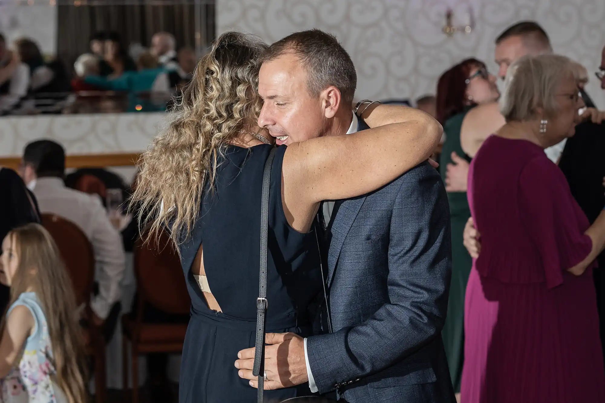 A man in a suit and a woman in a black dress with wavy blonde hair embrace and dance at a formal event. Other guests, including adults and a child, are mingling or dancing in the softly lit background.