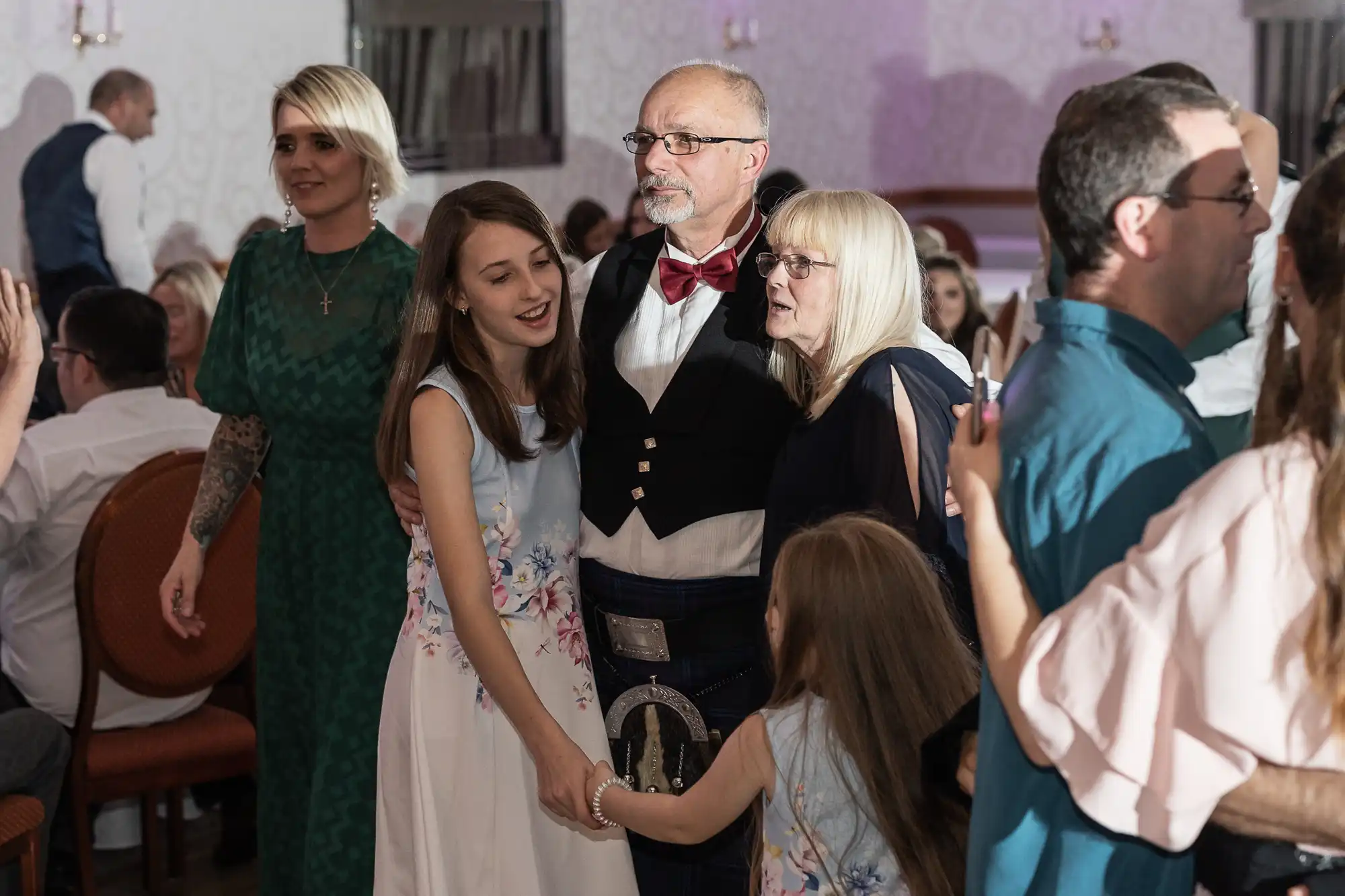 A group of people, including two girls in white floral dresses, a man in a kilt and bow tie, and a woman with blonde hair and glasses, stand together smiling and embracing at an indoor event with others seated and mingling in the background.