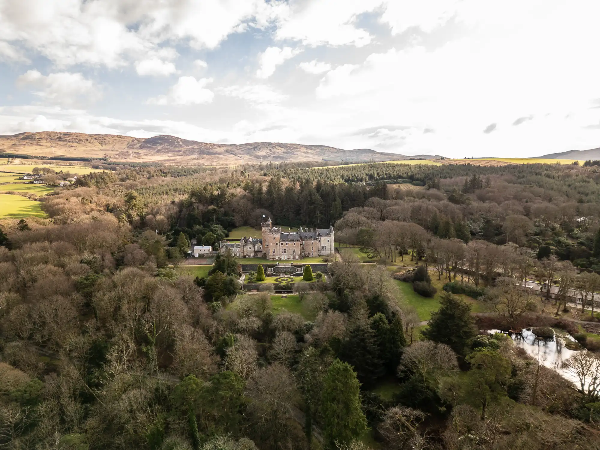 A large stone mansion, Glenapp Castle, with turrets and elaborate gardens sits surrounded by dense trees; rolling hills and fields stretch into the background under a mostly cloudy sky. A reflective pond is visible near the trees in the lower right corner.