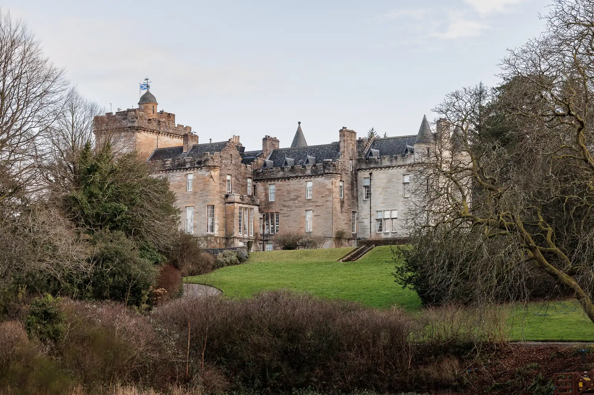 A large historic stone castle with battlements and turrets stands on a green lawn, surrounded by leafless trees and shrubs under a pale blue sky. The castle’s multiple chimneys and tall windows are visible, giving it a stately, mediaeval appearance.