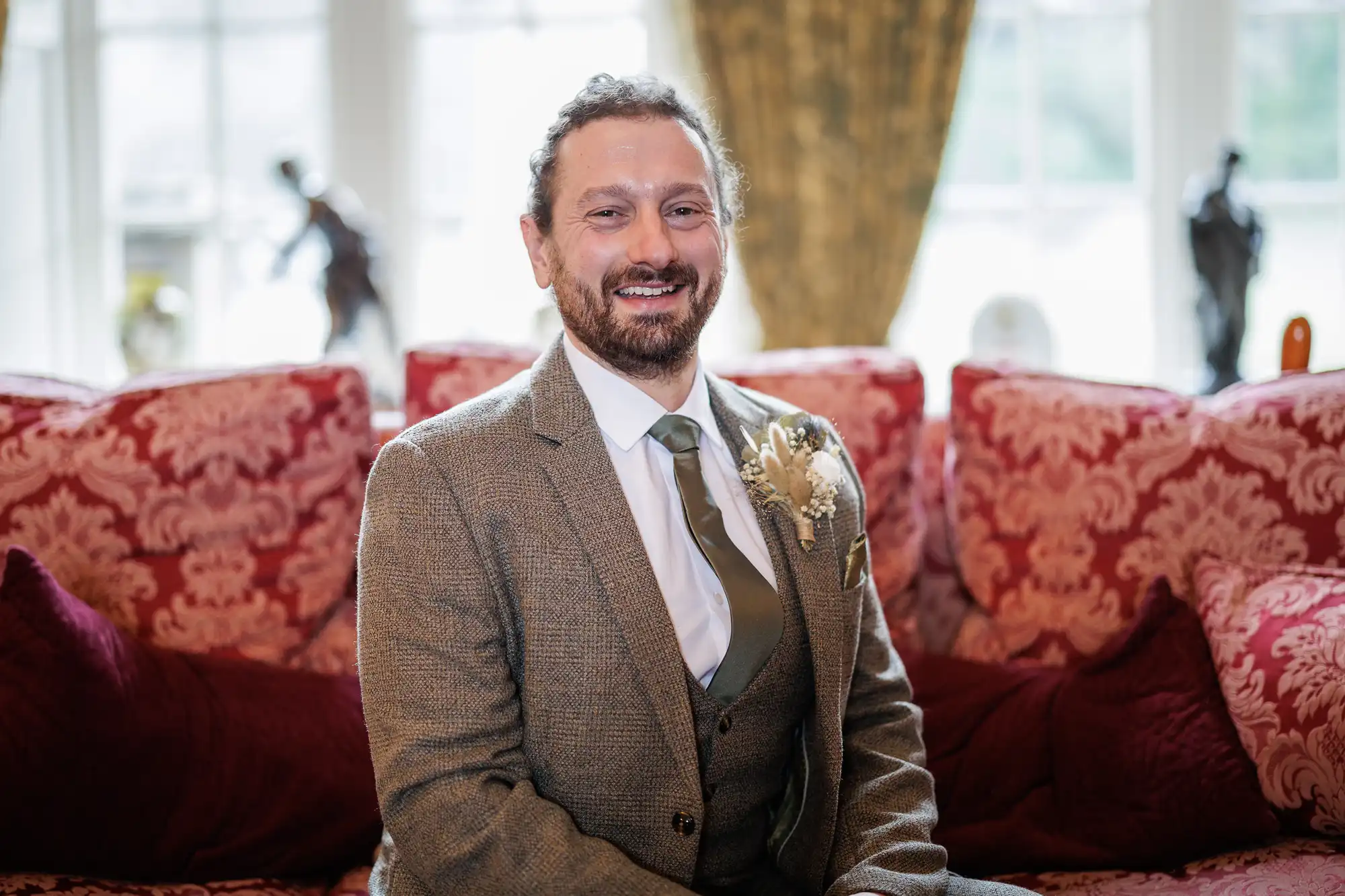 A man with curly dark hair and a beard sits on a red patterned sofa, smiling. He wears a brown tweed suit, white shirt, and green tie with a floral buttonhole. Ornate cushions and a window with statues and gold curtains are in the background.