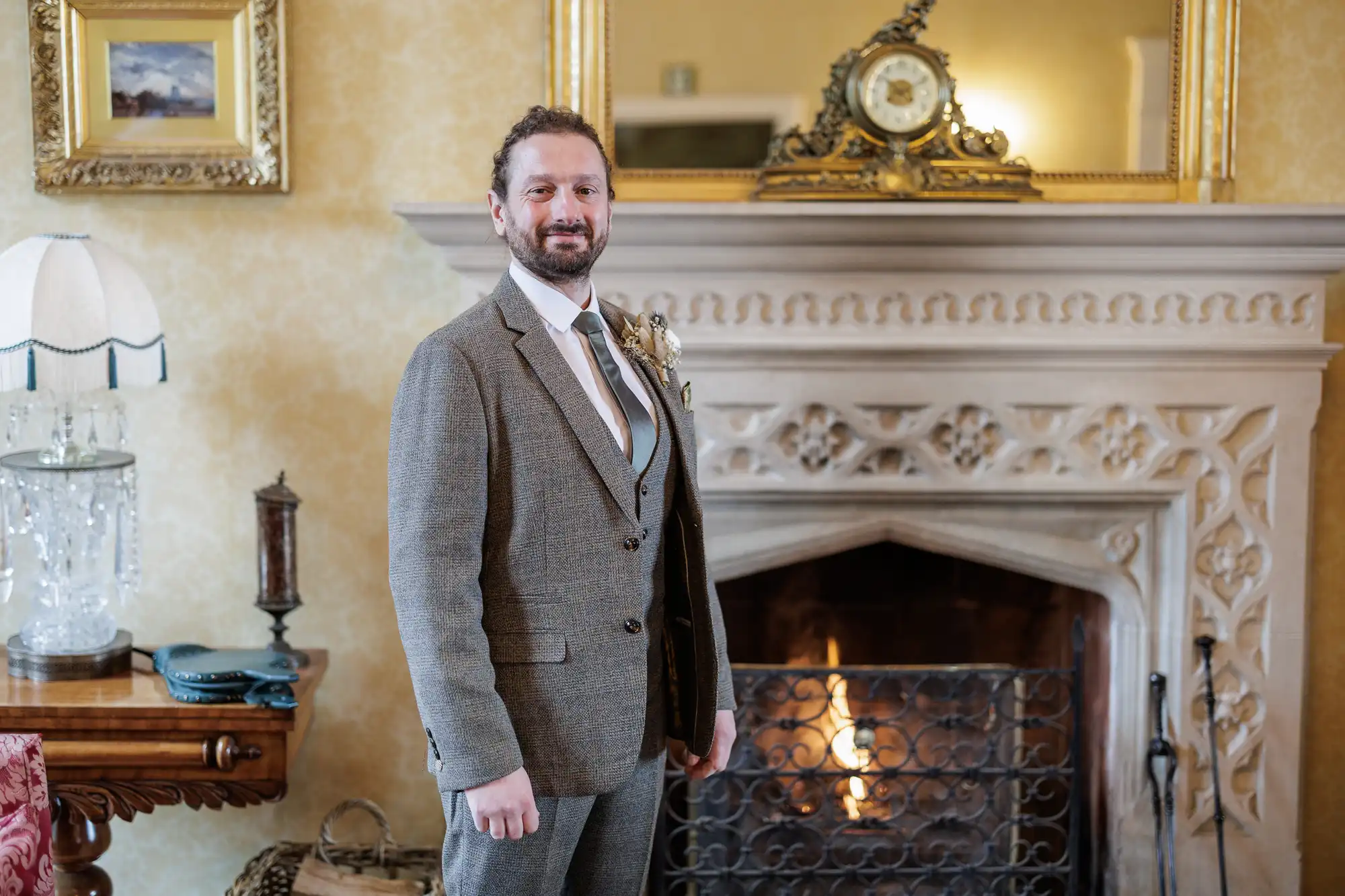A man in a grey suit and tie stands smiling in front of an ornate fireplace with a lit fire. The room has elegant décor, including a large mirror, framed art, and antique furnishings. The atmosphere is warm and sophisticated.