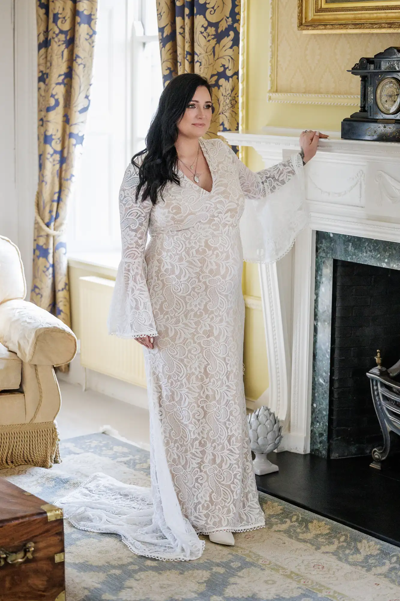 A woman with long dark hair stands indoors by a fireplace, wearing a white lace gown with bell sleeves and a train. She has a calm expression, holding the mantelpiece with one hand. The room has ornate decor, patterned curtains, and vintage furnishings.