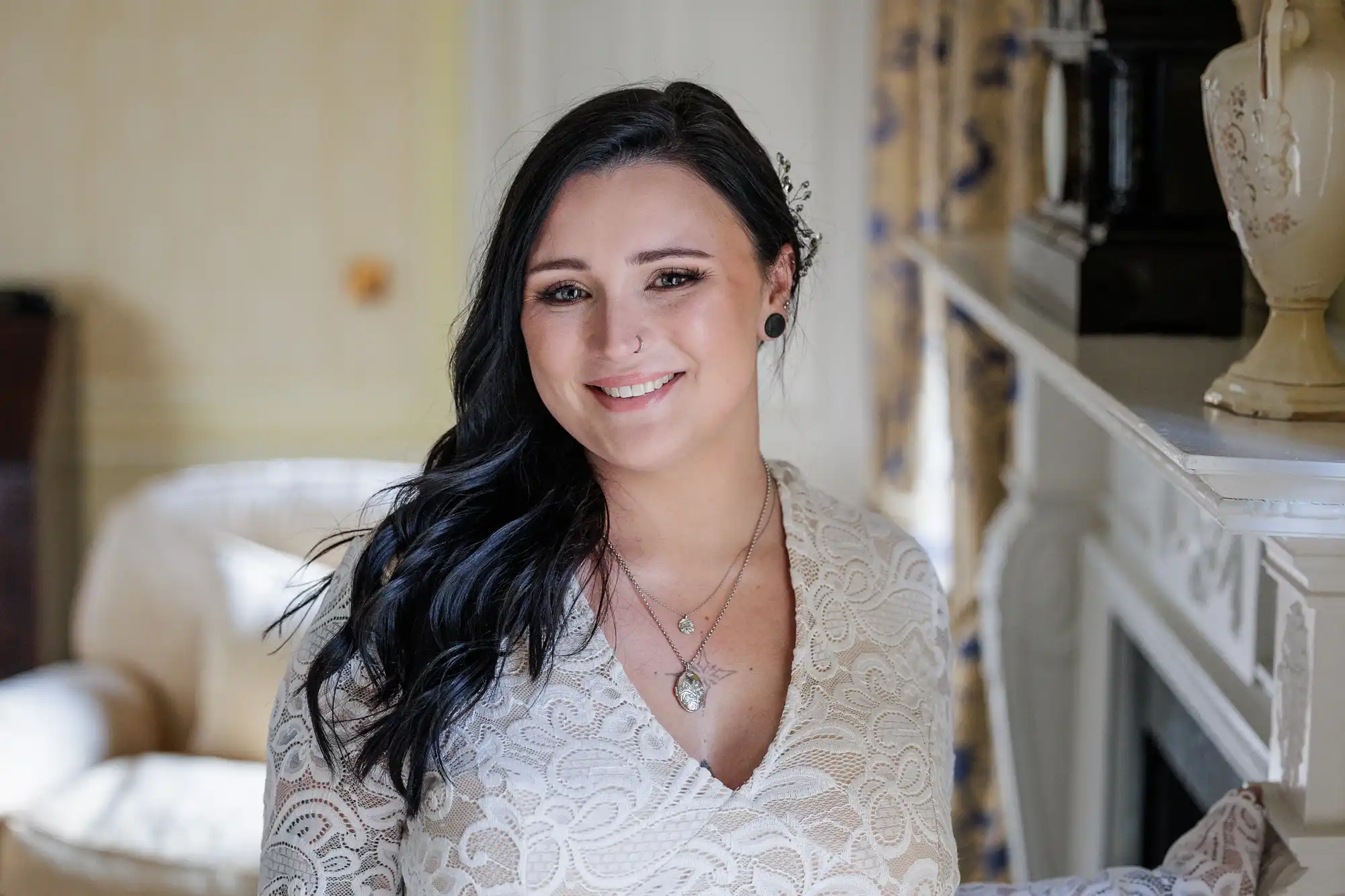 A woman with long dark hair, wearing a cream lace dress and layered necklaces, smiles at the camera. She stands indoors by a white fireplace in a softly lit room with cream and yellow tones and floral curtains in the background.
