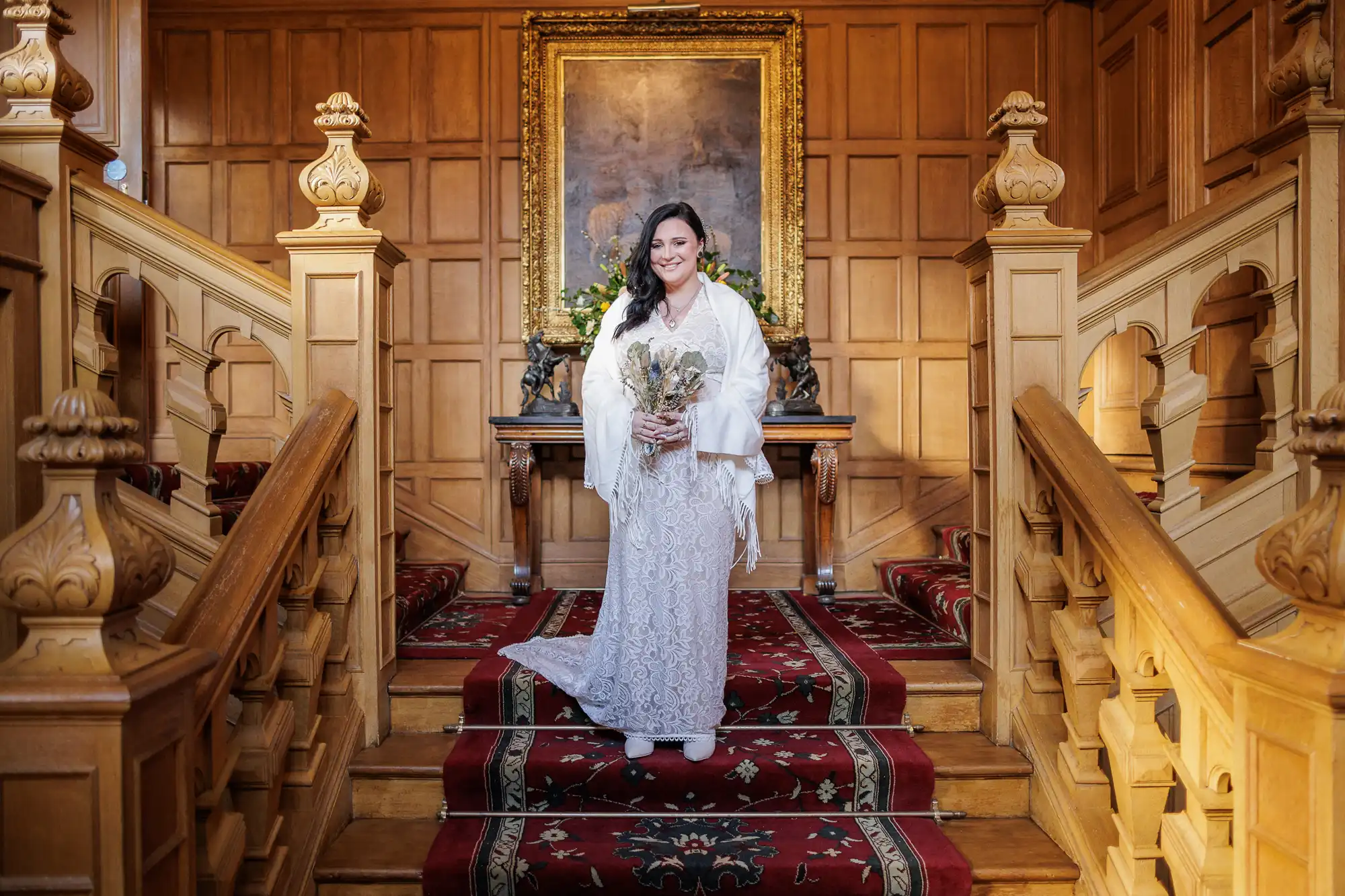 A bride in a white lace dress stands smiling on a red-carpeted staircase with ornate wooden railings. She holds a bouquet of dried flowers. Behind her, a large framed painting hangs on a wood-panelled wall above a table with decorations.