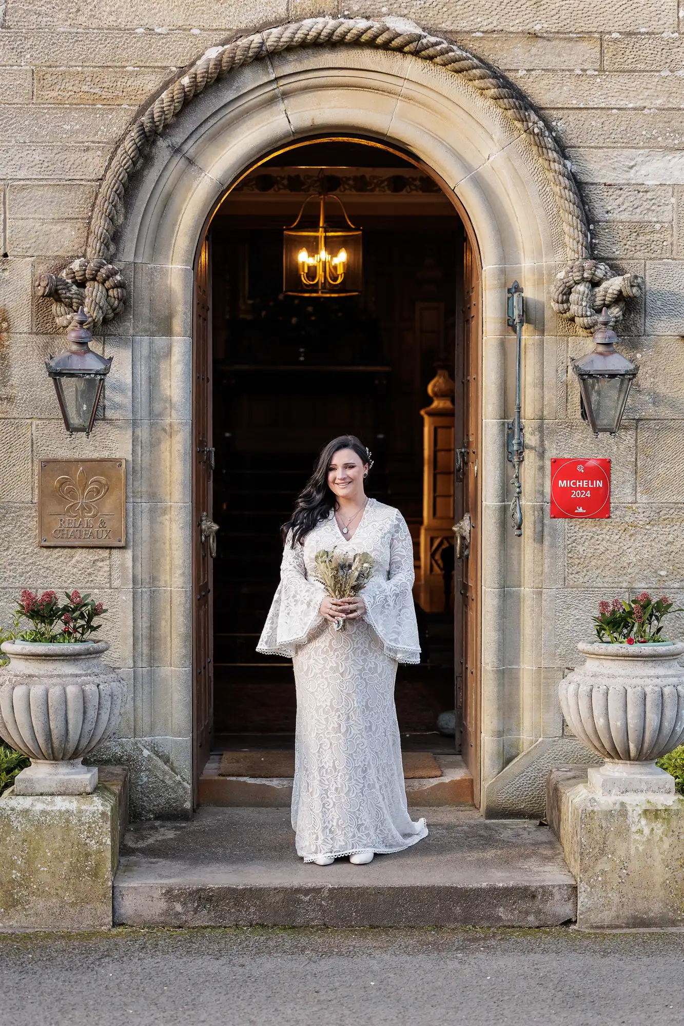 A smiling bride in a long white lace dress stands in a stone doorway beneath a rope arch. She holds a bouquet and looks forward. The doorway is framed by two potted plants and wall lanterns; a red Michelin plaque is visible on the right wall.