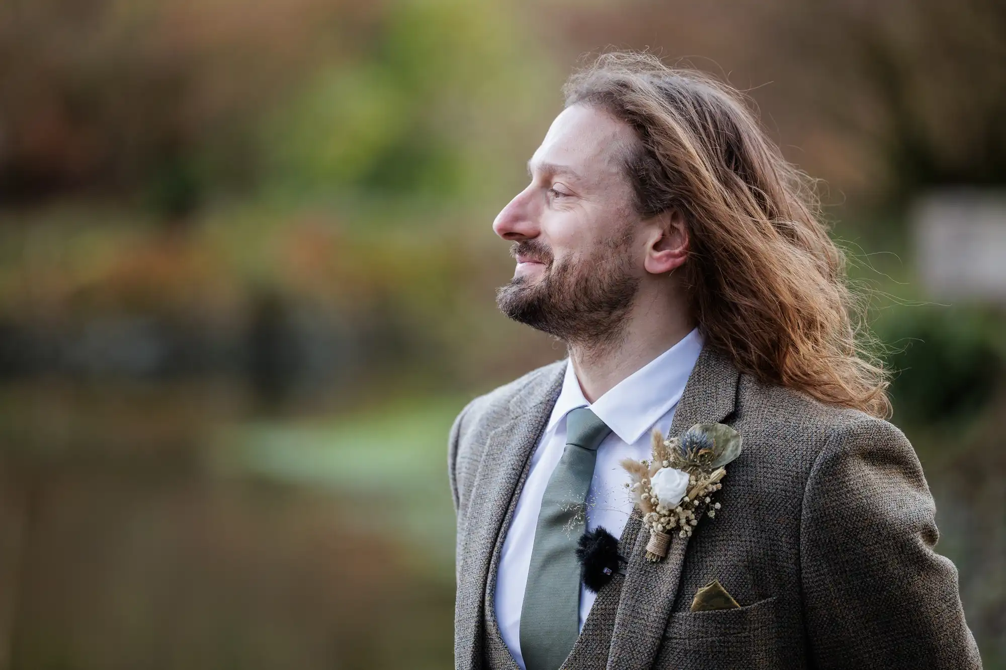 A man with long brown hair and a beard stands outdoors, smiling and looking to the right. He wears a grey tweed suit, white shirt, green tie, and a buttonhole with white flowers and dried plants. The background is blurred greenery.
