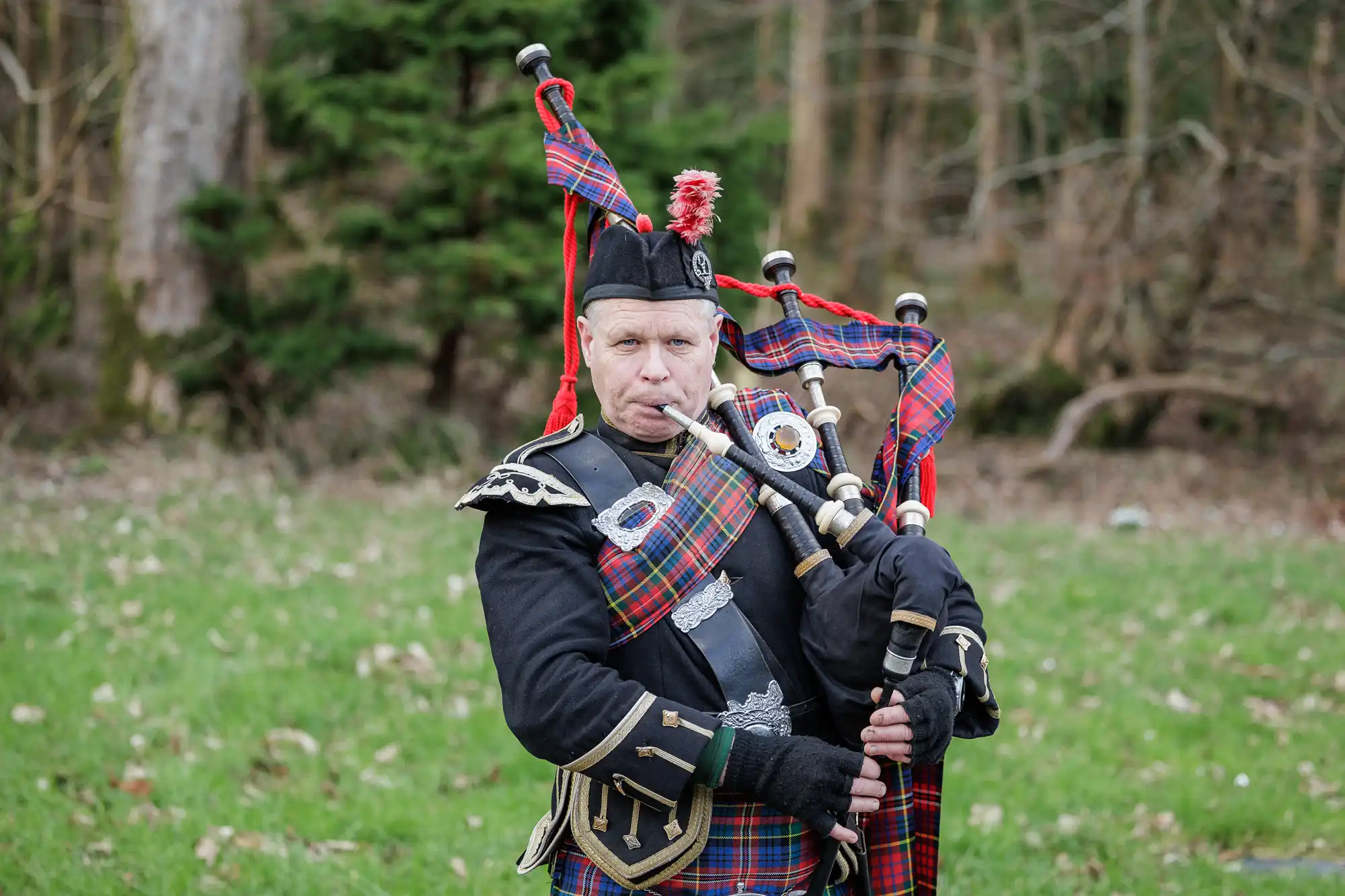 A man in traditional Scottish attire plays bagpipes outdoors. He wears a tartan-patterned sash, black jacket with silver details, black hat with a red pompom, and stands on grass with trees and shrubs in the background.
