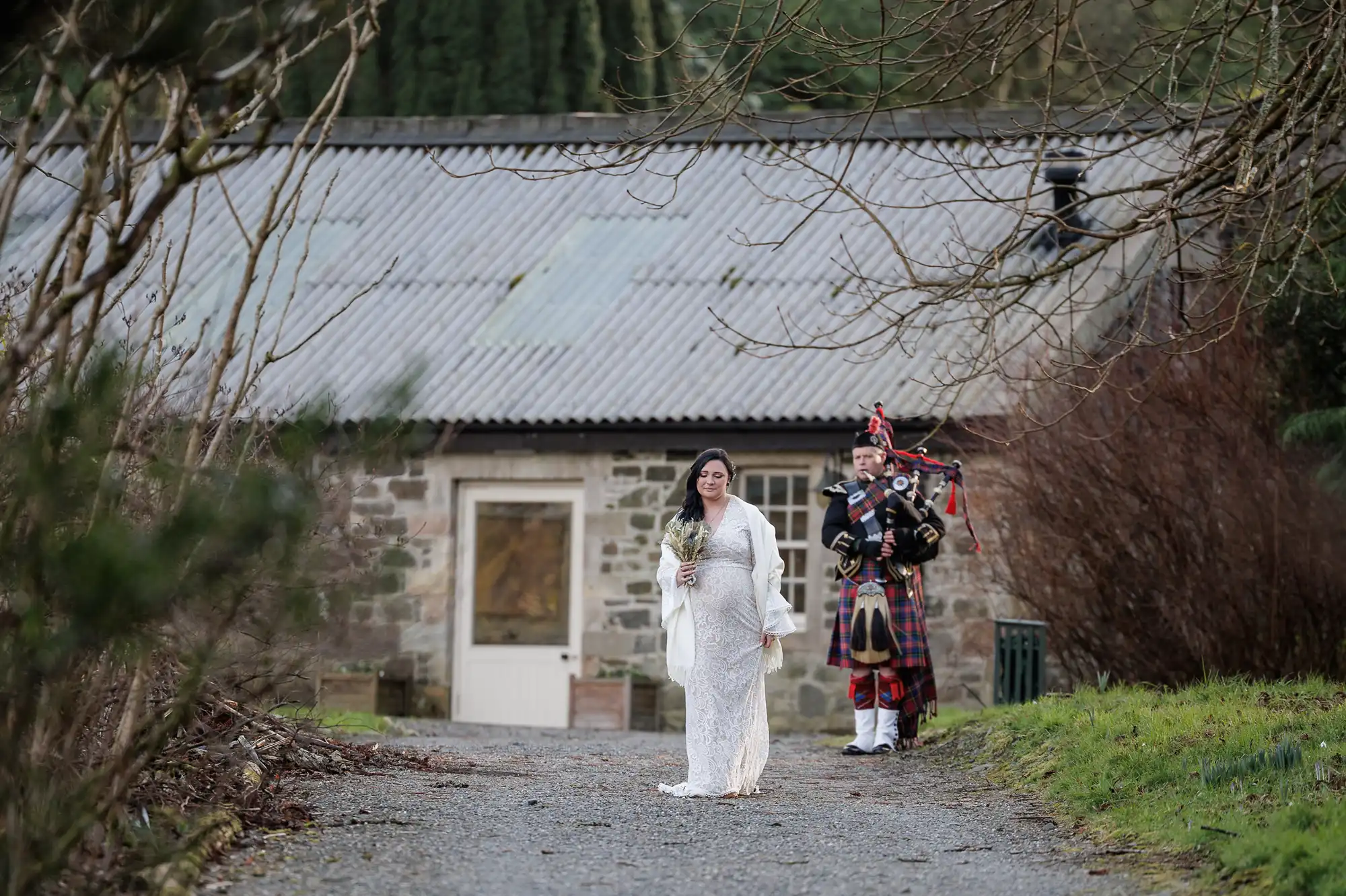 A bride in a white dress walks on a gravel path holding a bouquet, followed by a bagpiper in traditional Scottish attire. They are outside near a rustic stone building with a corrugated roof, surrounded by bare trees and greenery.