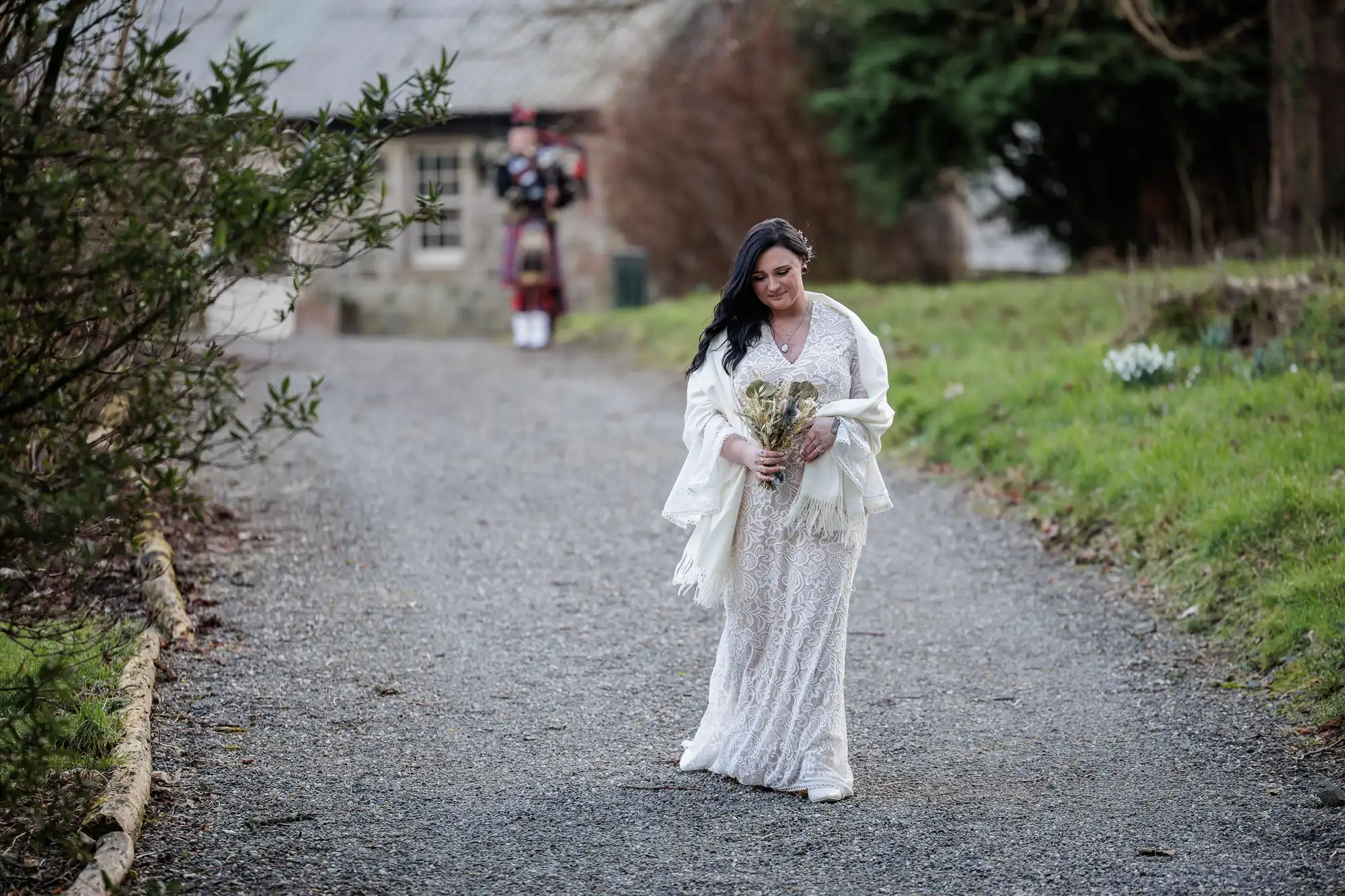 A bride in a white lace dress and shawl walks alone on a gravel path, holding a bouquet. Green grass and trees line the sides. In the background, a bagpiper in traditional Scottish attire stands near a building. The scene appears peaceful and natural.