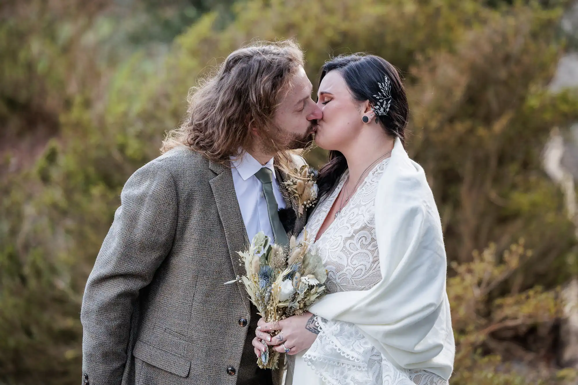 A couple kisses outdoors at their wedding. The groom wears a grey suit and has long, wavy hair; the bride wears a white lace dress with a shawl, holding a bouquet of dried flowers. Green foliage fills the softly blurred background.
