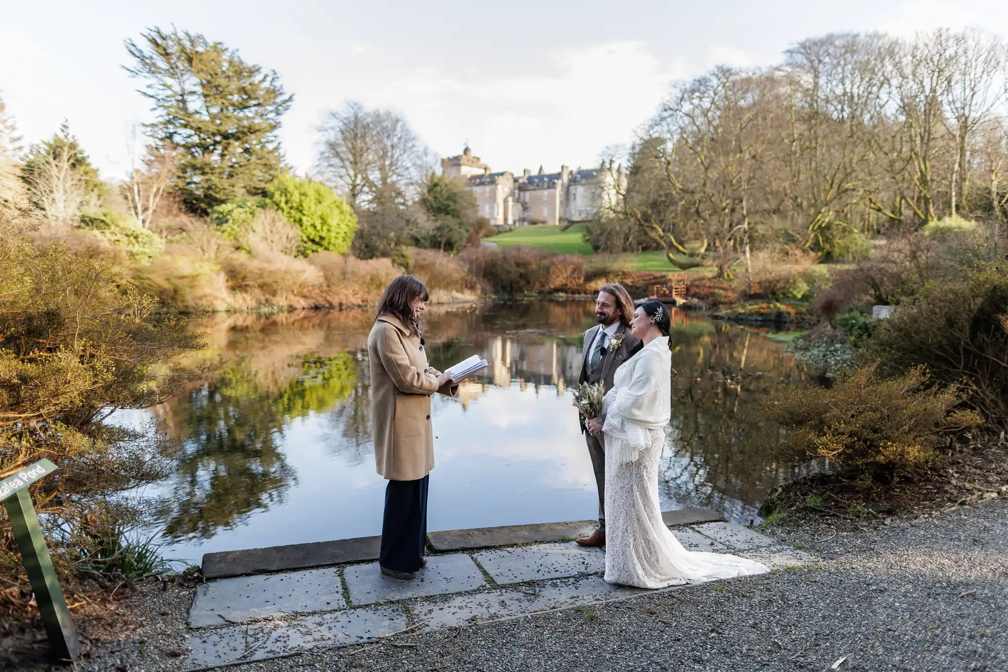 A bride and groom stand by a calm pond, facing an officiant who reads from a book. The bride wears a white gown, the groom a tan suit. Behind them, trees surround the water, and a large castle sits atop a hill in the distance under a bright sky.
