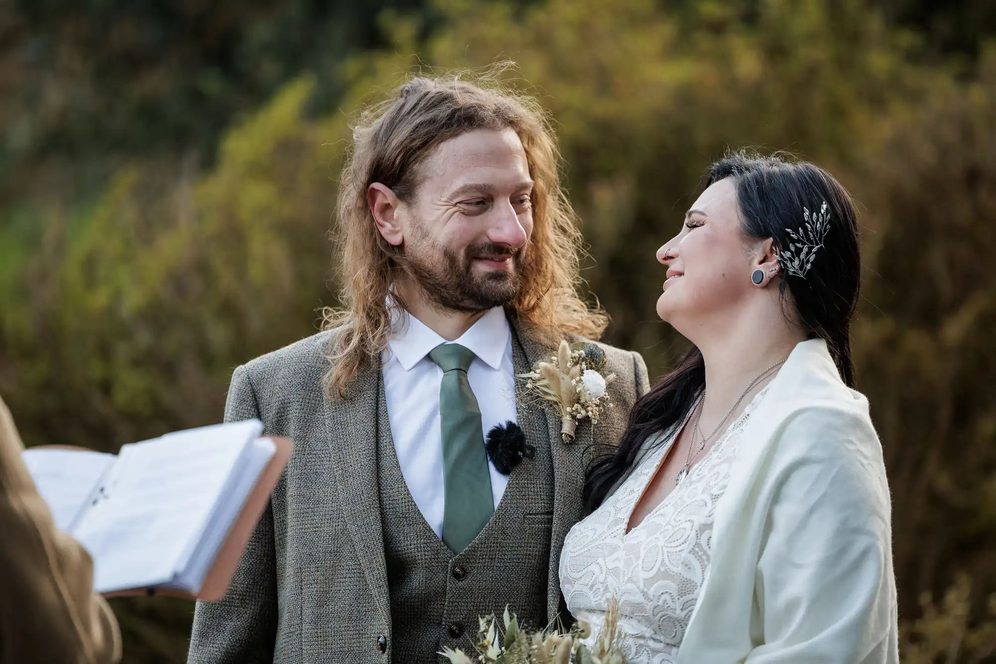 A couple stands close together outdoors, gazing at each other and smiling. The man has long, wavy brown hair, a beard, and wears a brown suit with a white shirt and green tie. The woman has long dark hair, a hair accessory, and wears a white dress with a shawl.