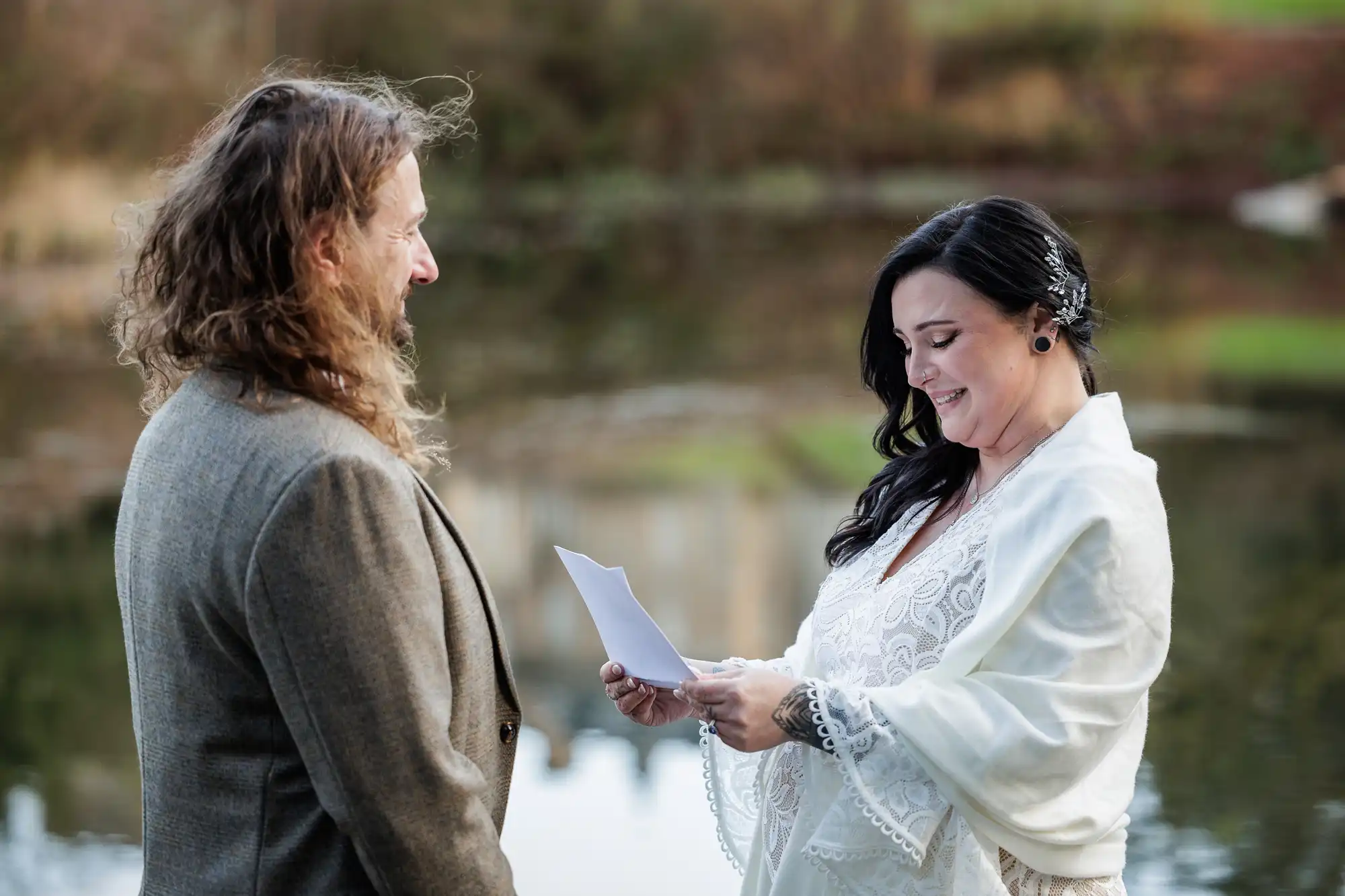A woman in a white dress and shawl smiles whilst reading from a piece of paper, standing outdoors by a pond, facing a man with long brown hair and a beard in a brown suit jacket. The background is blurred, showing grass and water.