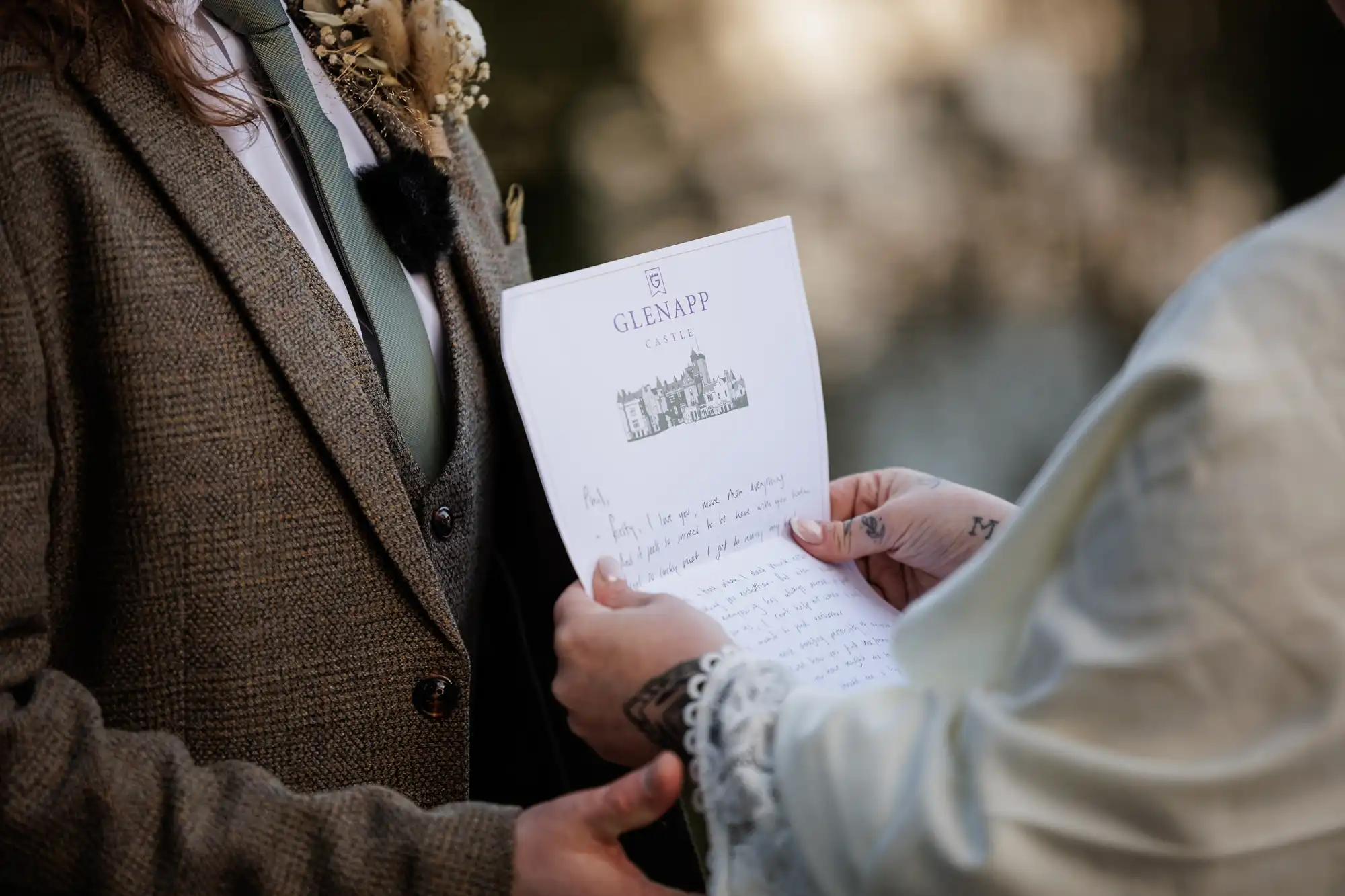 A person in a brown tweed suit with a green tie stands facing another person, who holds a handwritten letter on Glenapp Castle stationery. Only their torsos and hands are visible, with soft outdoor lighting and blurred greenery in the background.