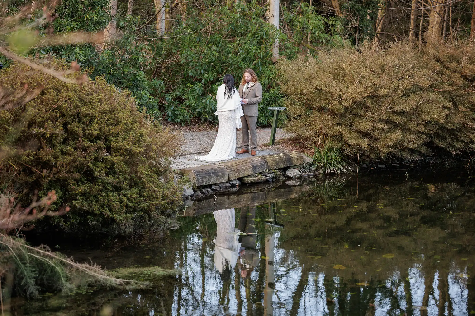 A bride and groom stand facing each other on a small jetty beside a pond, surrounded by dense bushes and trees. Their reflections are visible in the water. The scene is calm and intimate, set in natural, wooded surroundings.
