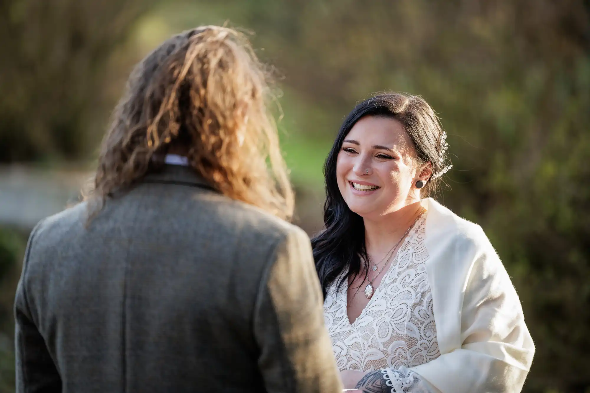 A smiling bride with long dark hair, wearing a lace dress and white shawl, faces a person with long wavy hair in a brown jacket. They are outdoors, surrounded by greenery, sharing a joyful moment. The sunlight highlights the bride’s happy expression.