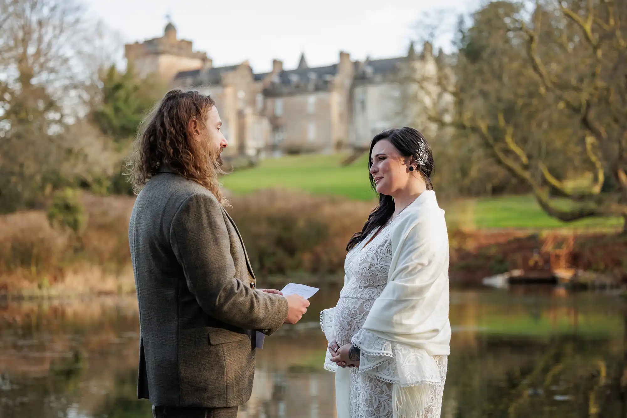 A couple stands by a calm lake, facing each other. The man holds papers and wears a brown suit; the woman wears a white lace dress and shawl. Behind them, there is green grass, trees, and a large historic building under a clear sky.