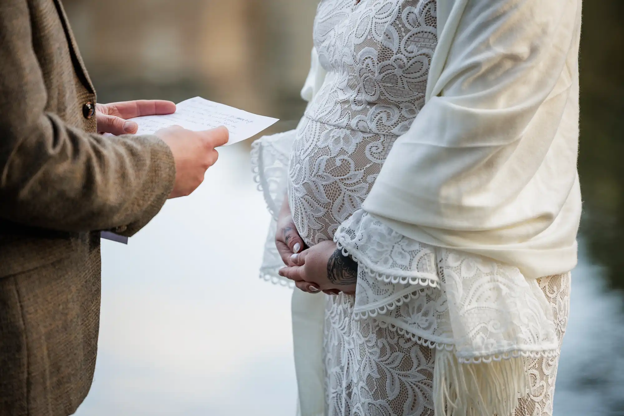 A close-up of a pregnant person in a white lace dress with a shawl, holding their belly, standing next to someone in a brown jacket who is reading from a handwritten note. The background is softly blurred, suggesting an outdoor setting near water.