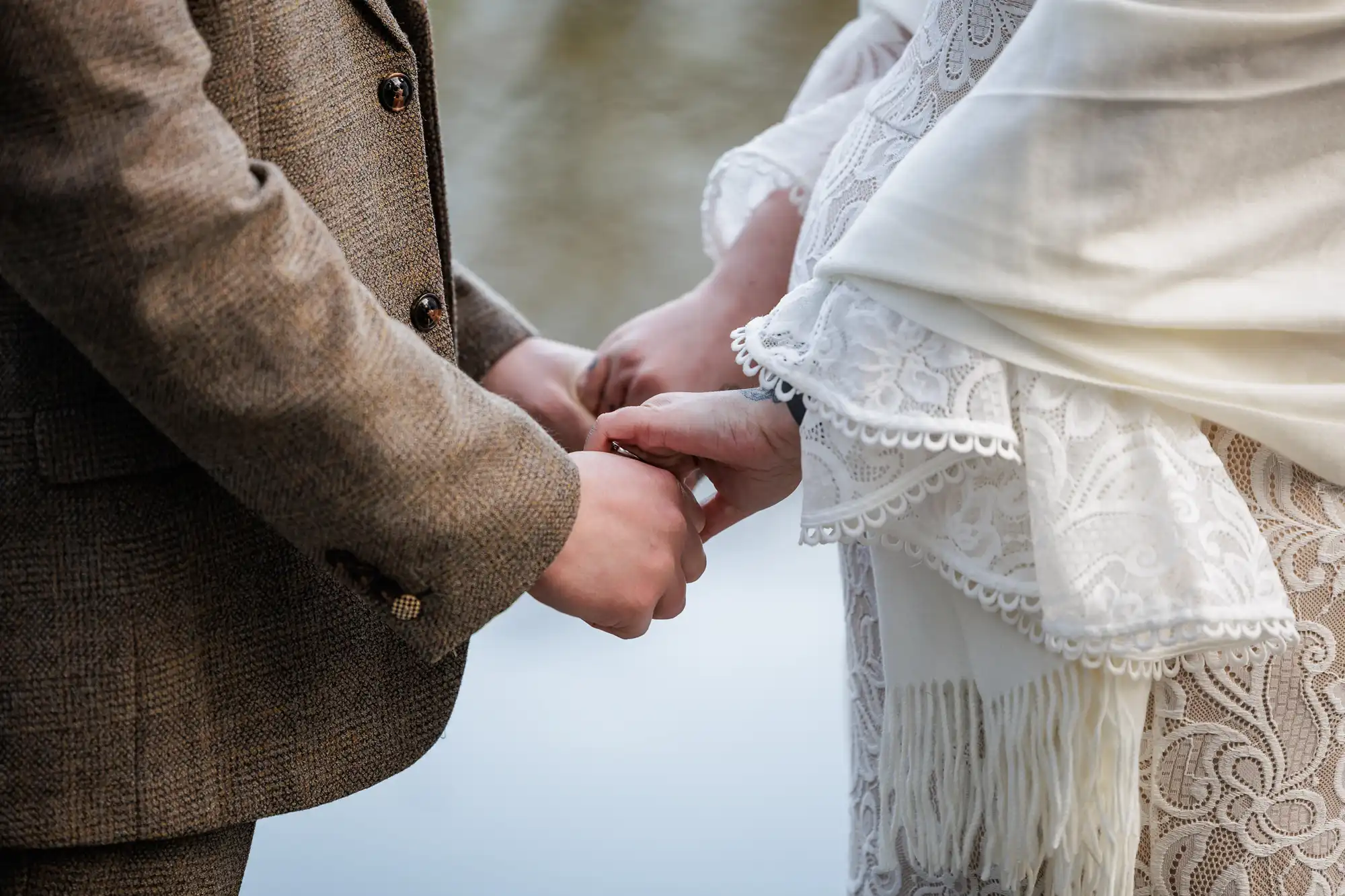 Close-up of a couple holding hands outdoors. One person wears a brown tweed suit; the other wears a white lace dress with a shawl. The background is blurred, suggesting water. The moment appears intimate and romantic, likely from a wedding or engagement.