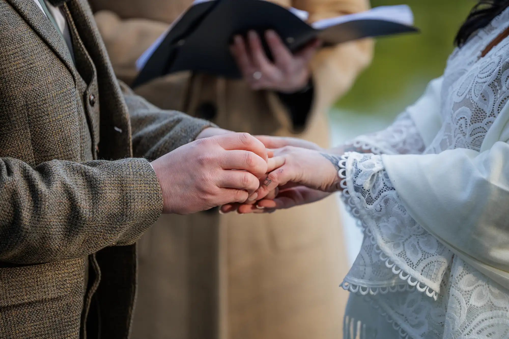 Close-up of two people holding hands during a wedding ceremony. One person wears a brown suit, the other a white lacy dress. An officiant holding an open book stands in the blurred background. The scene is outdoors.
