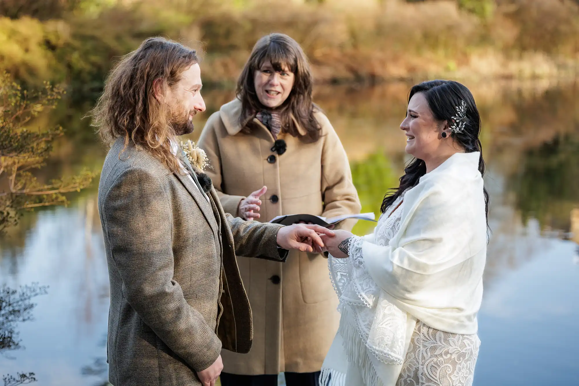 A couple exchange rings during an outdoor wedding by a lake, smiling at each other. The groom wears a tweed suit; the bride wears a white lace shawl. An officiant in a tan coat stands between them. Trees and water are in the blurred background.