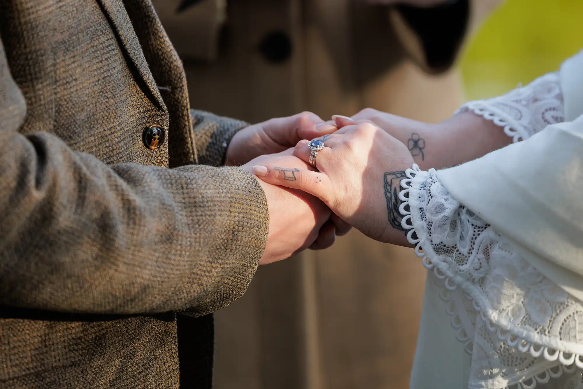 Two people stand close, holding hands. One wears a brown tweed jacket; the other wears a white lace sleeve and has visible tattoos and a ring with a blue stone. They appear to be in an intimate or ceremonial moment, with blurred greenery in the background.