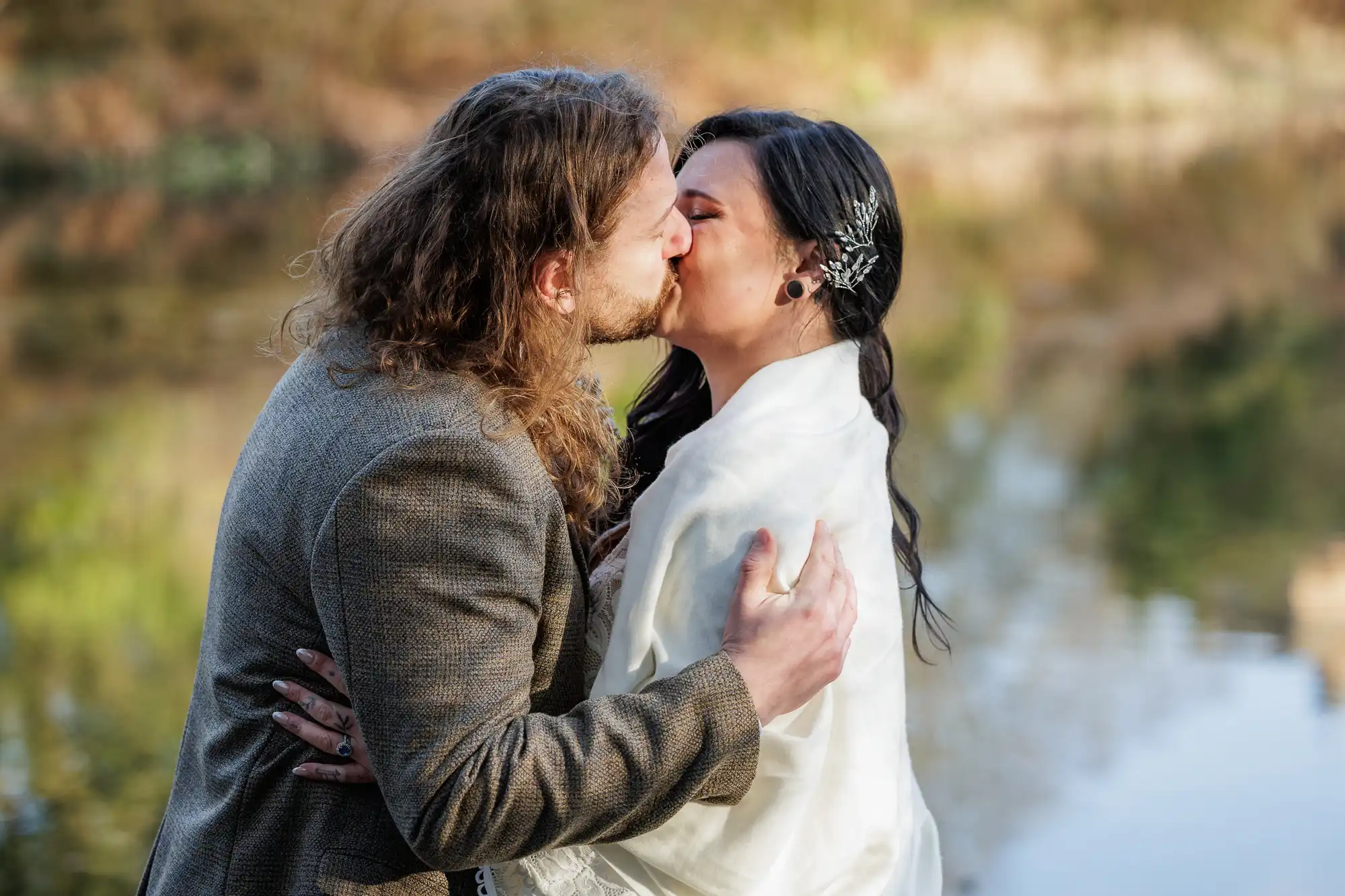 A couple share a kiss outdoors by a blurred lake. The man has long, wavy hair and wears a brown suit. The woman has dark hair adorned with a decorative hairpin and is wrapped in a white shawl. Both appear happy and affectionate.