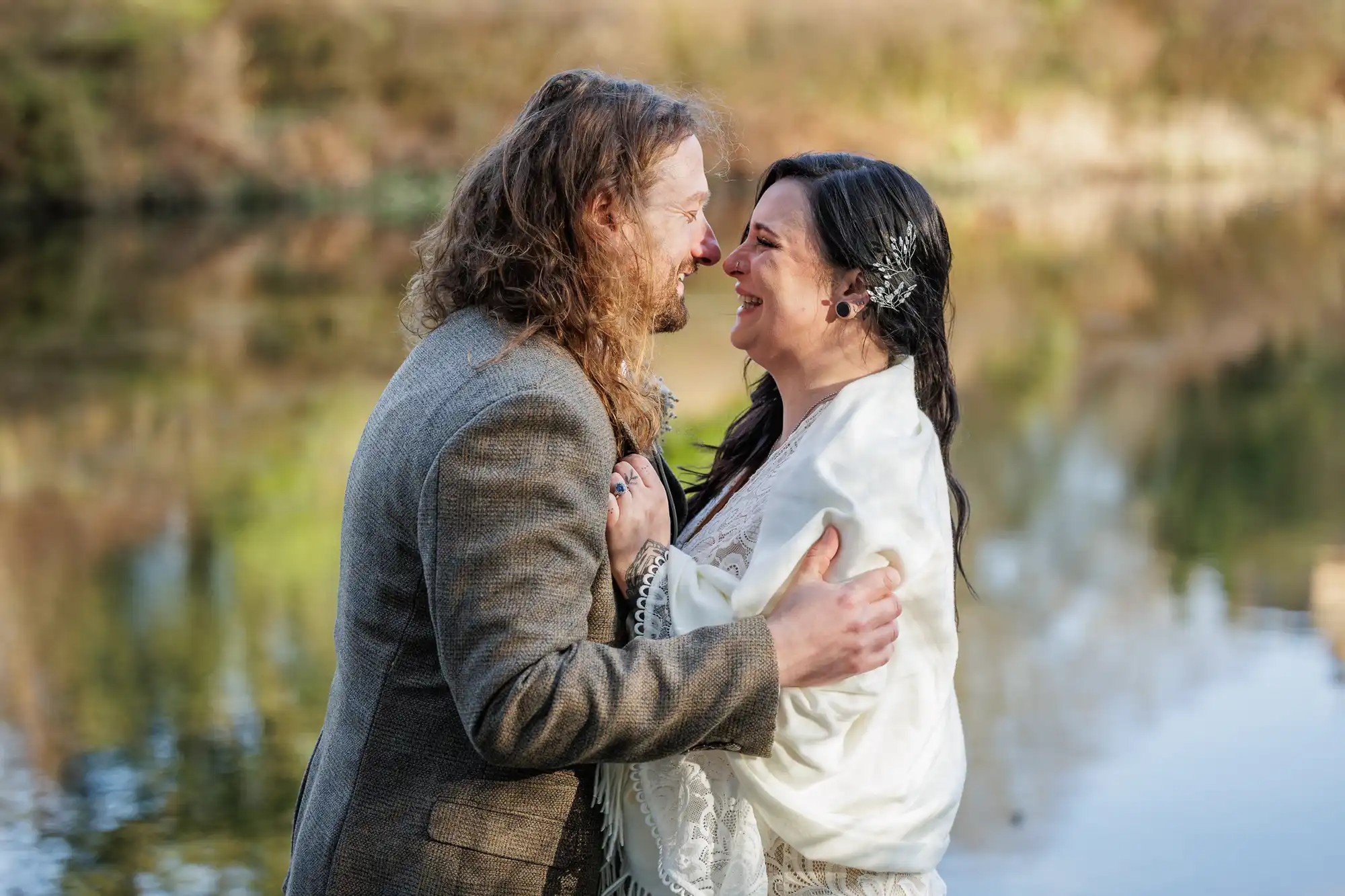 A smiling couple stands close together outdoors by a calm body of water, gazing into each other’s eyes. The man has long, wavy hair and wears a brown jacket, while the woman wears a white shawl and hair clip with decorative details.