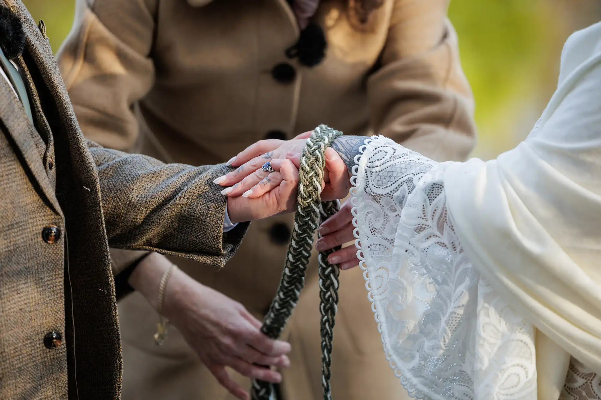 Three people participate in a handfasting ceremony. Two clasped hands, one in a lace sleeve and one in a suit, are wrapped with braided cords by a third person, symbolising unity and marriage. The background is softly blurred with greenery.