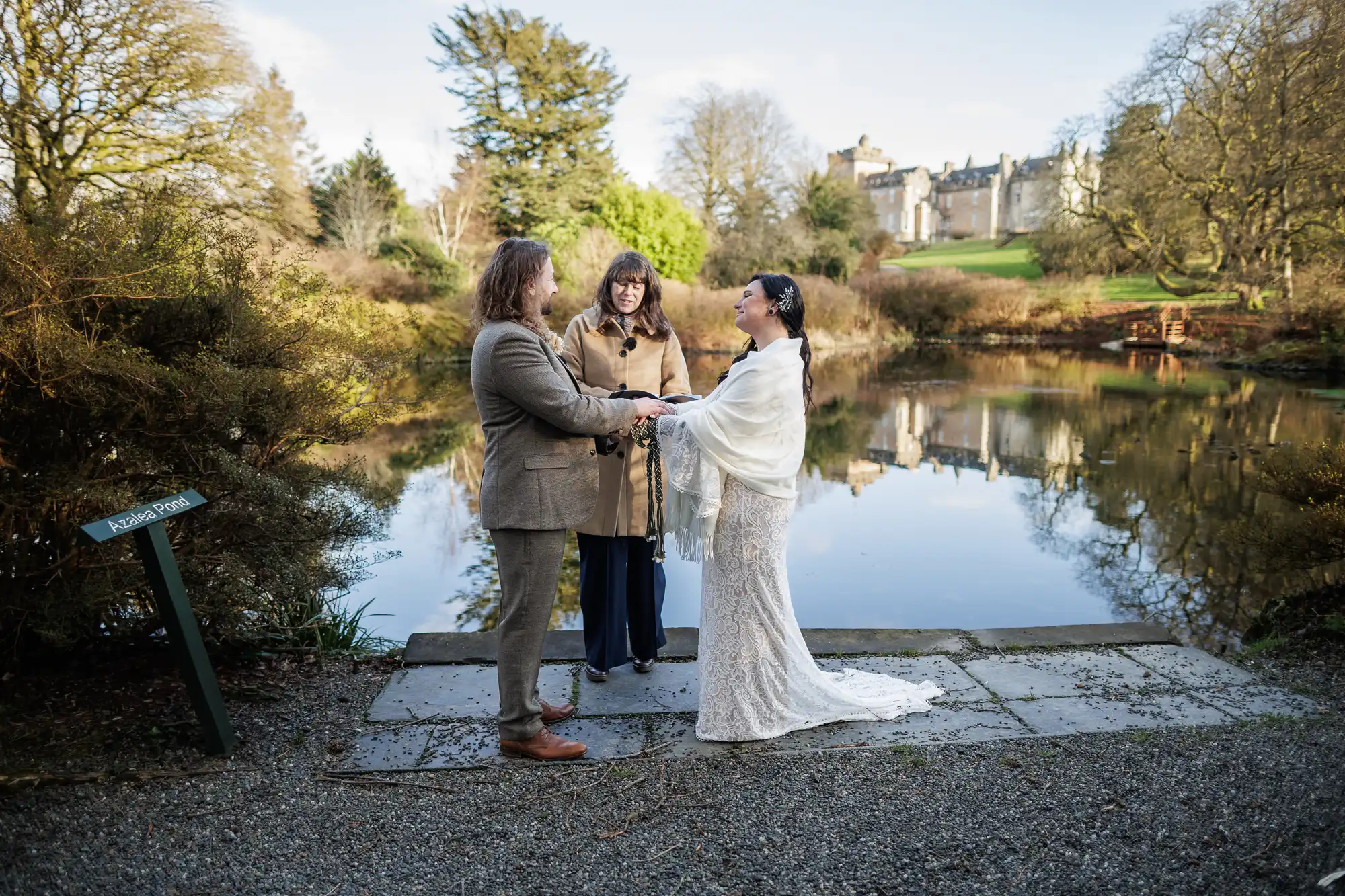 A couple stands facing each other, holding hands in wedding attire by a pond. An officiant stands between them. Leafless trees, greenery, and a castle are visible in the background under a clear sky. A sign reading “Kensington Park” is in the foreground.