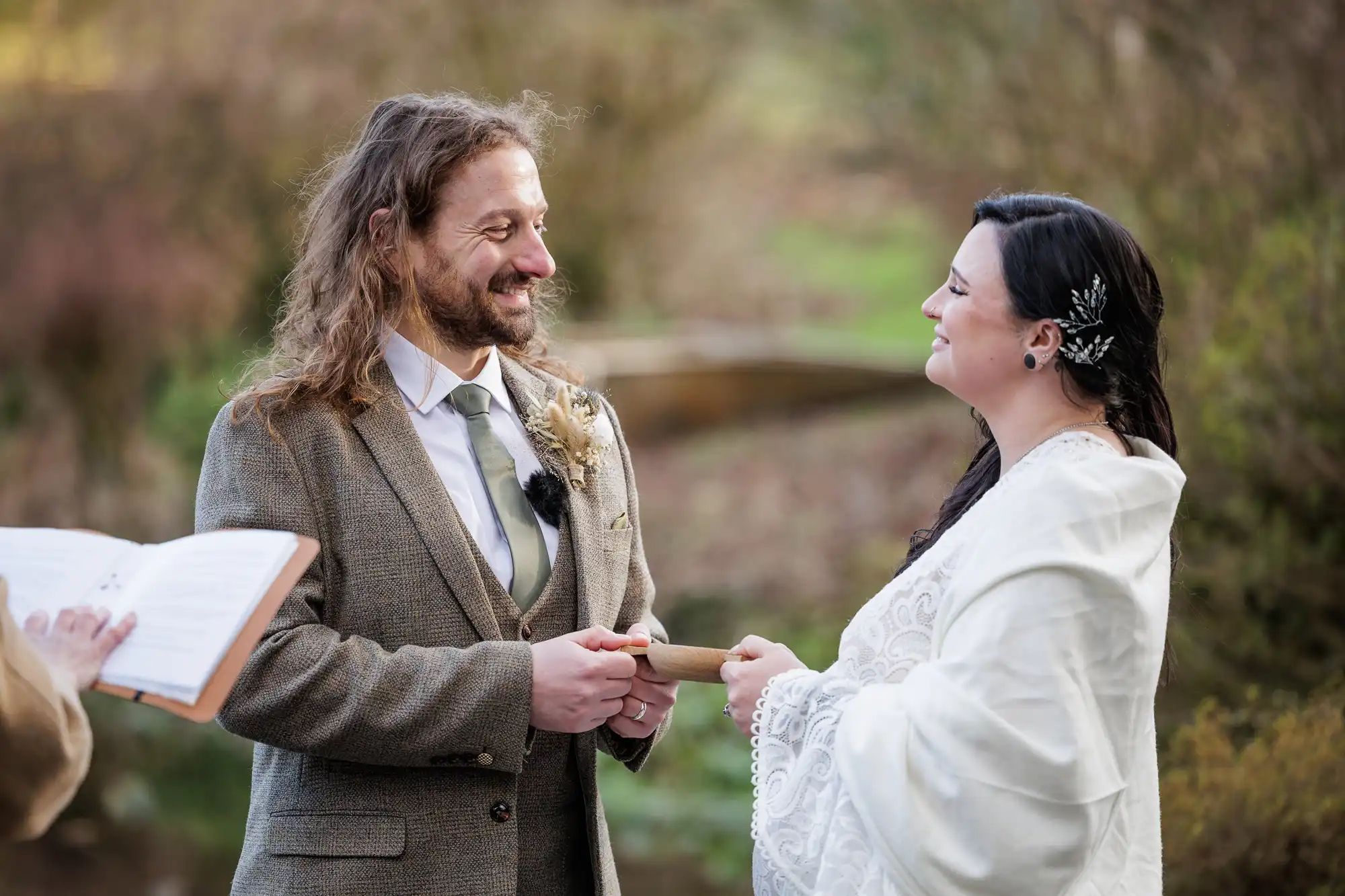 A man in a tweed suit and a woman in a white shawl and lace dress smile warmly at each other, holding hands during an outdoor wedding ceremony. The setting is natural with blurred greenery in the background and an officiant holding a book.