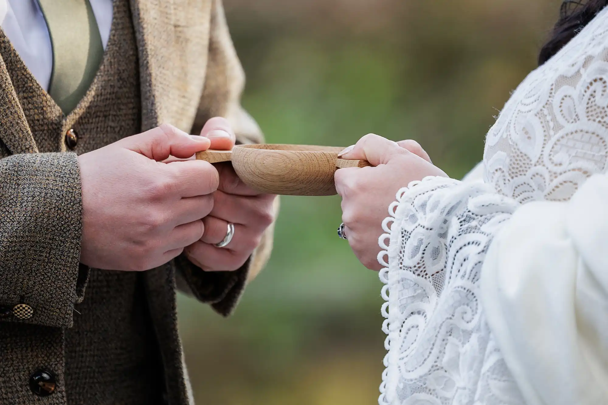 A close-up of two people in formal attire holding a shared wooden cup. One wears a tweed suit and tie, the other a white lace garment. Both hands meet at the cup, suggesting a ceremonial or symbolic exchange. Background is softly blurred.