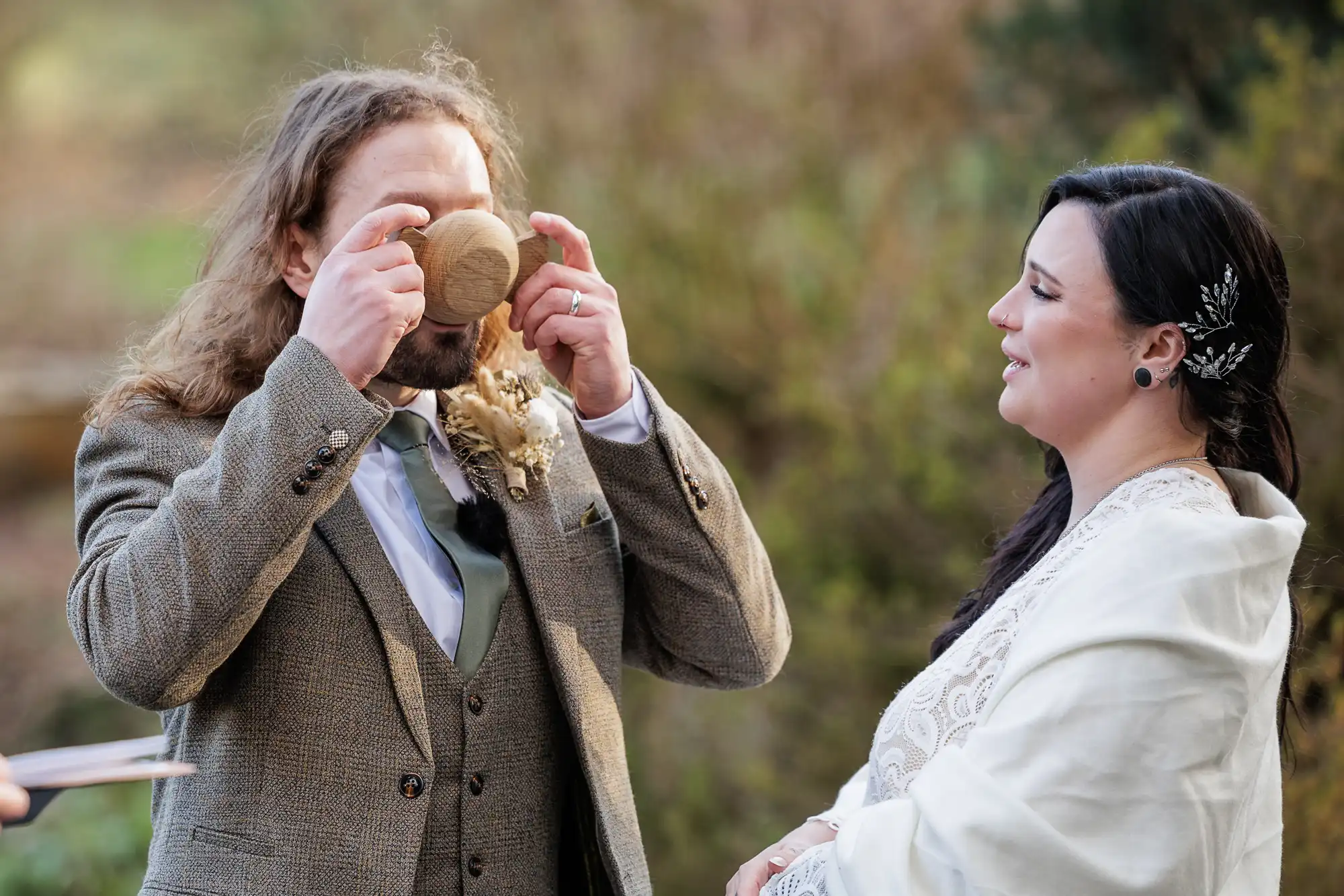 A man with long hair and a beard, wearing a tweed suit, drinks from a wooden cup during an outdoor ceremony. A woman in a white shawl and a lacy dress stands beside him, smiling, with trees and greenery in the blurred background.