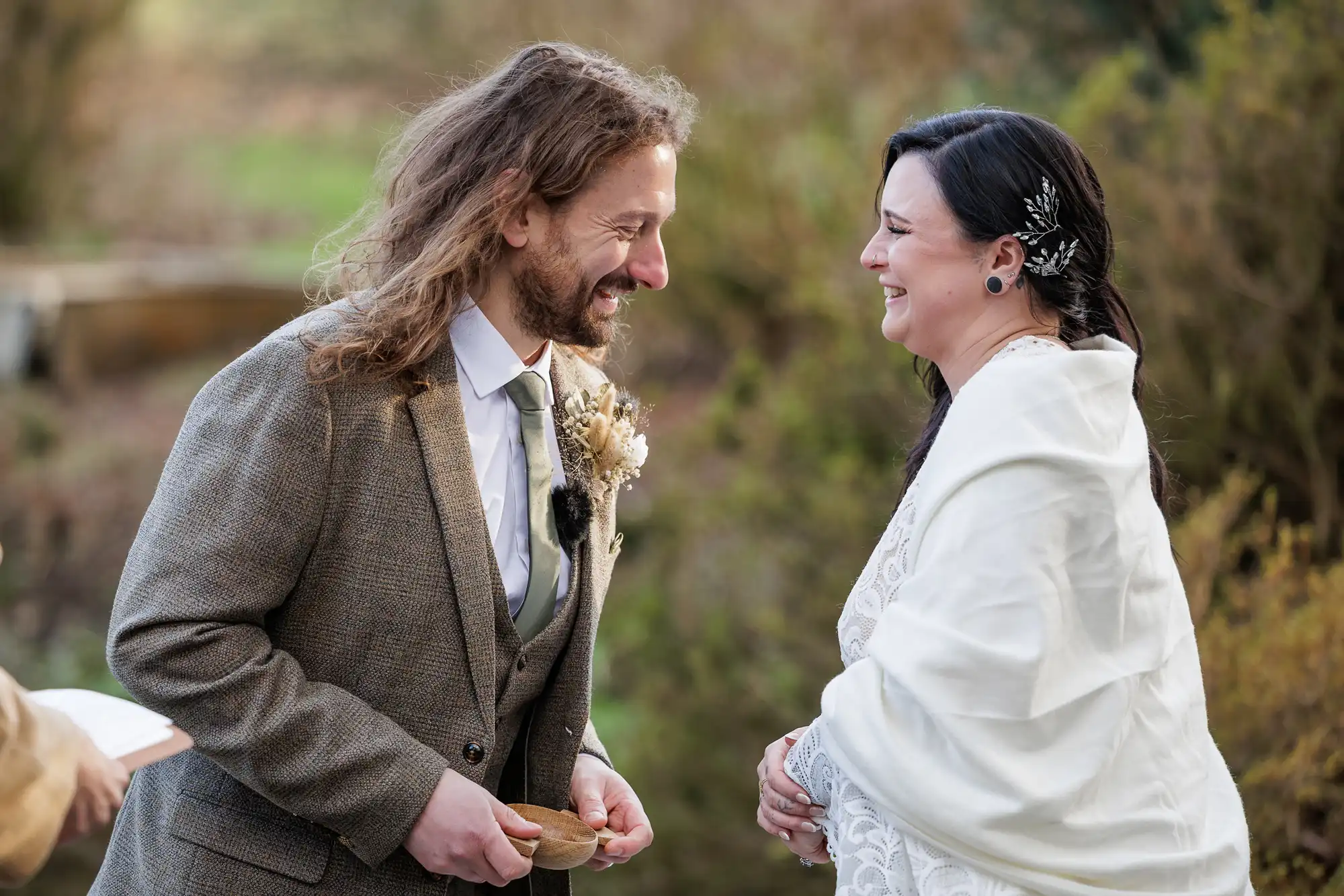 A man in a brown tweed suit and a woman in a white dress and wrap smile warmly at each other outdoors during a wedding ceremony, surrounded by greenery. The man holds a small bowl and the woman wears a decorative hair clip.