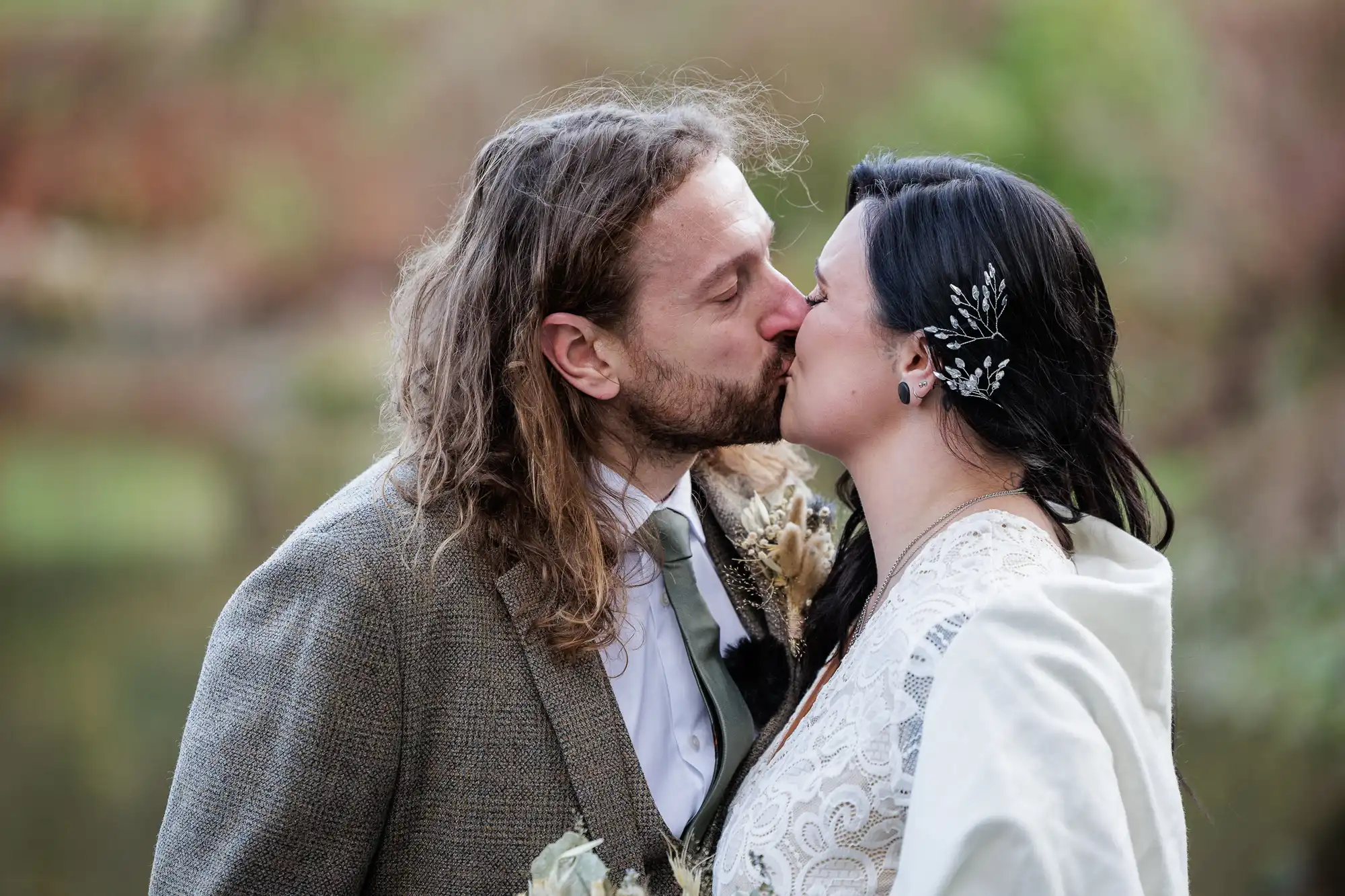 A man with long brown hair and a beard kisses a woman with black hair and a silver hair accessory. Both wear formal attire; he wears a tweed suit, she wears a white dress with lace. They stand outdoors, surrounded by soft greenery.