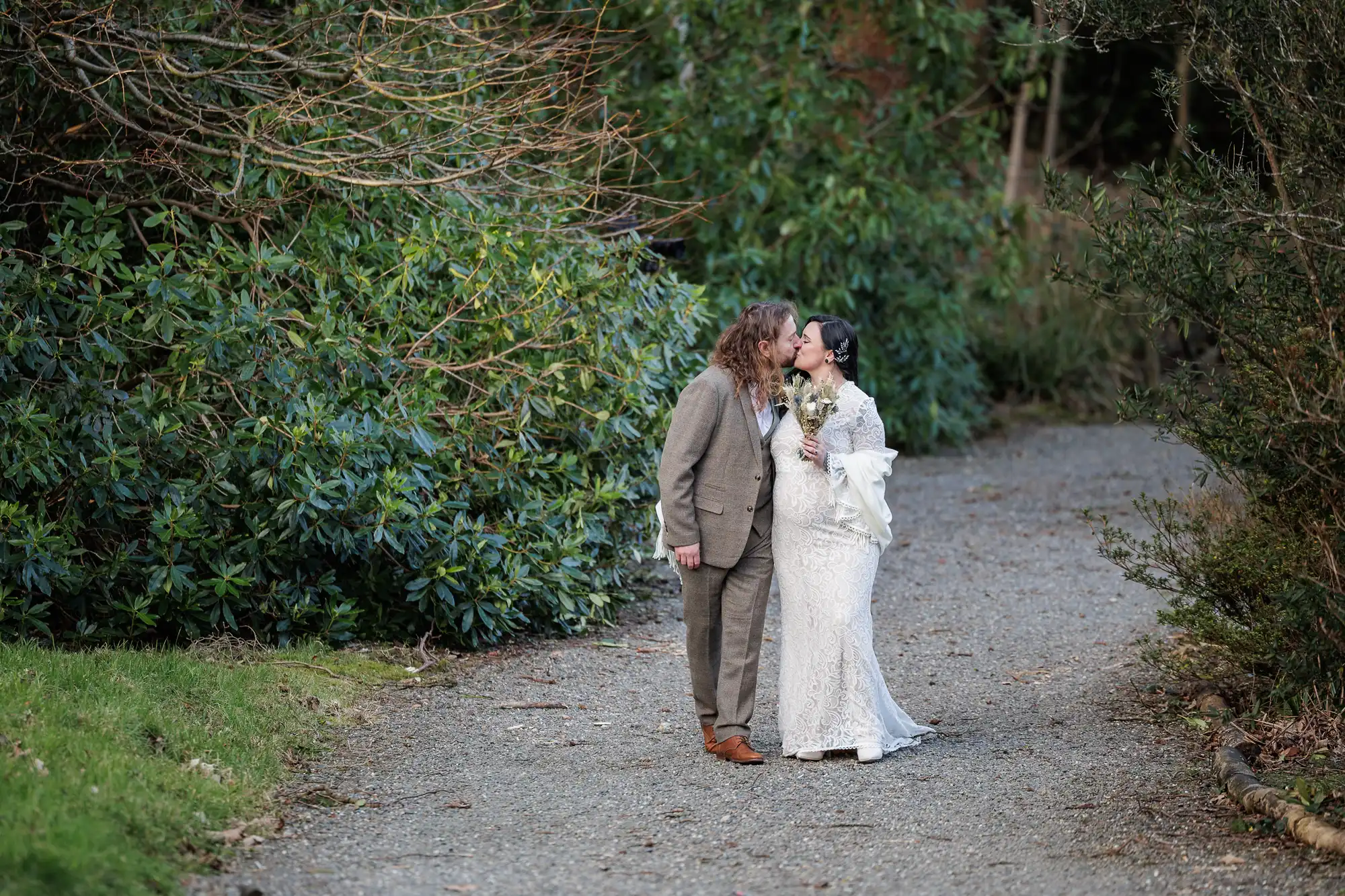 A couple dressed in wedding attire kiss on a gravel path surrounded by green, leafy trees and bushes. The groom wears a tweed suit, while the bride wears a white lace gown and holds a bouquet. They are outdoors, with natural greenery in the background.