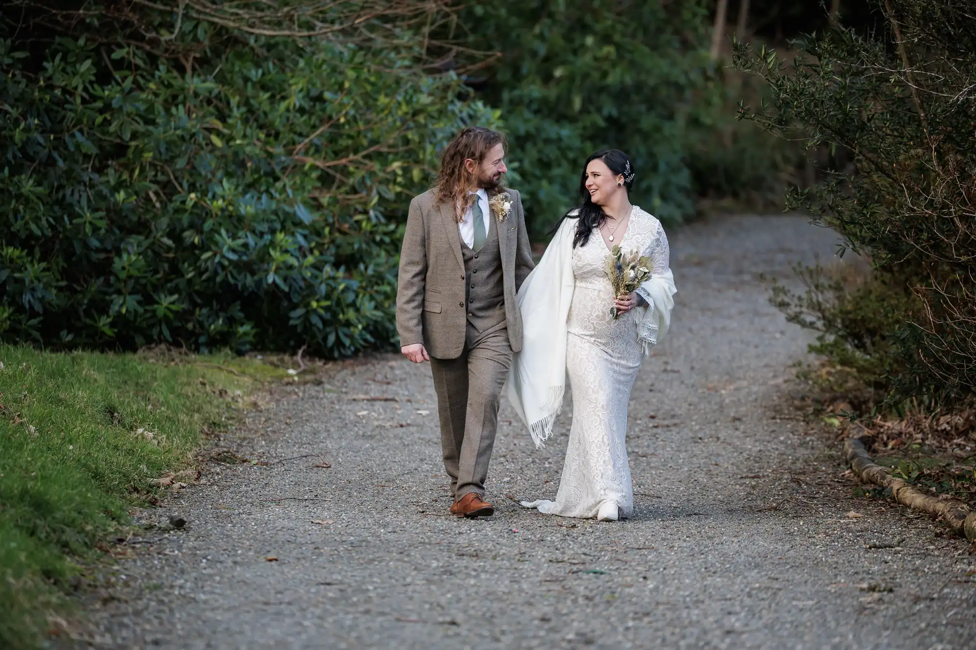 A bride in a white lace gown and shawl walks arm-in-arm with a groom in a brown suit on a wooded path. They smile at each other, holding a bouquet and enjoying a serene outdoor moment surrounded by greenery.