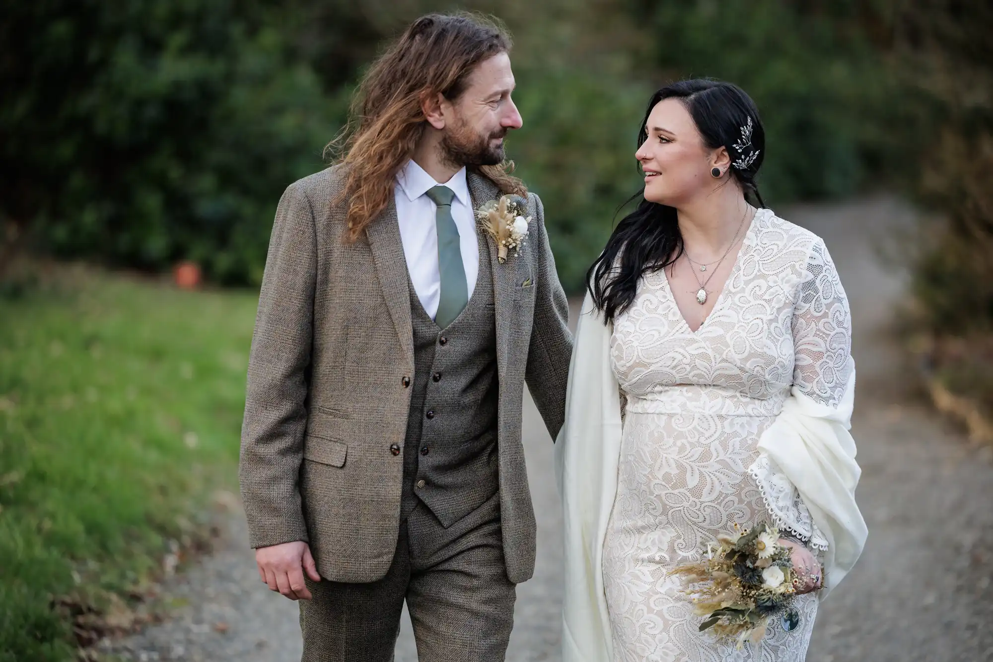A man in a brown suit and a woman in a lacy white dress with a shawl walk outside, smiling at each other. The woman holds a small bouquet of flowers. They appear happy, possibly newlyweds, with greenery and a gravel path in the background.